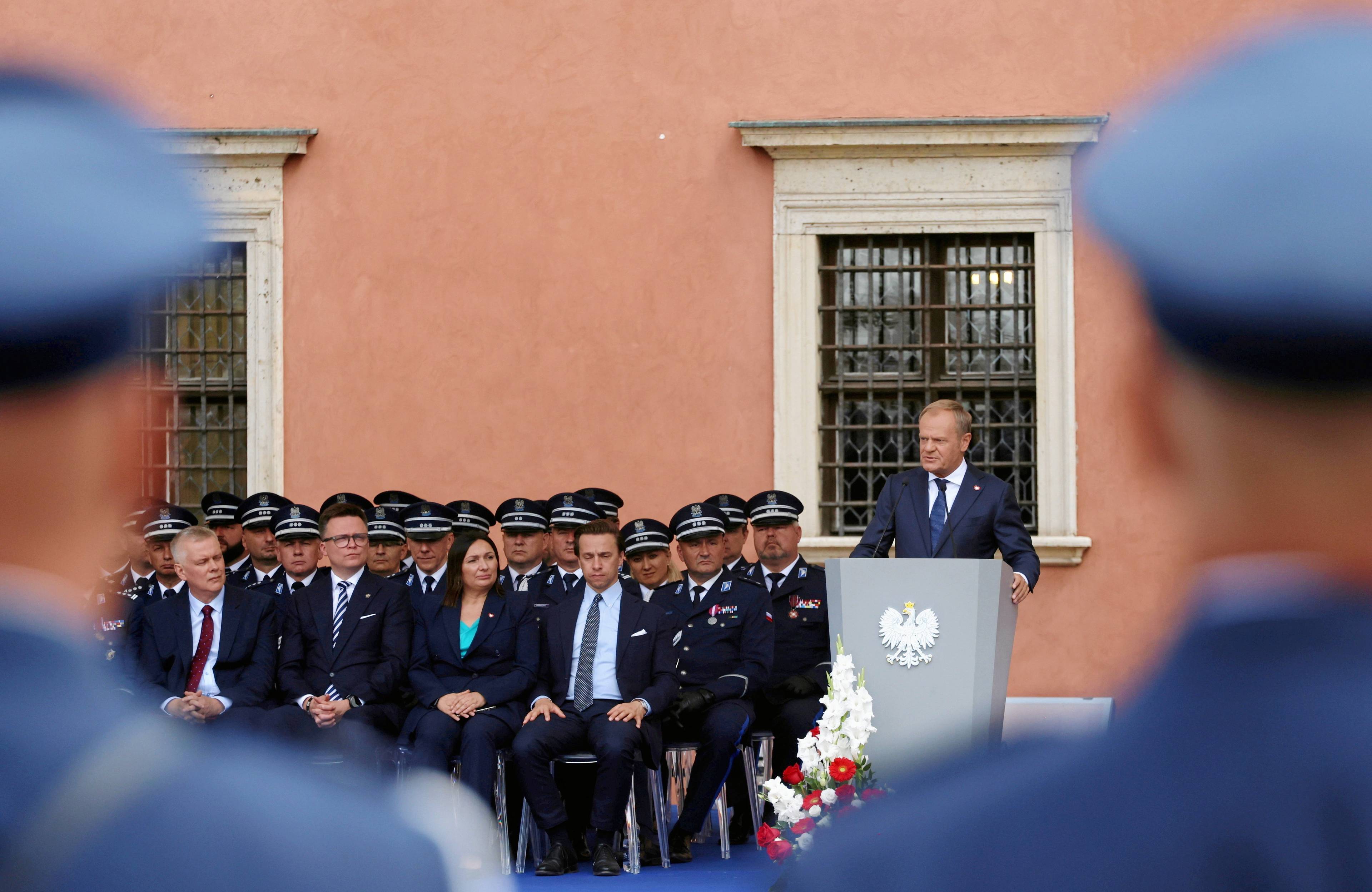 Mężczyzna w granatowym garniturze stoi za mównicą, obok niego trzy rzędy krzeseł, na których zasiadają oficjele w garniturach oraz policyjnych mundurach, na pierwszym planie zdjęcia rozmazany zarys dwóch mężczyzn w niebieskich mundurach i policyjnych czapkach.