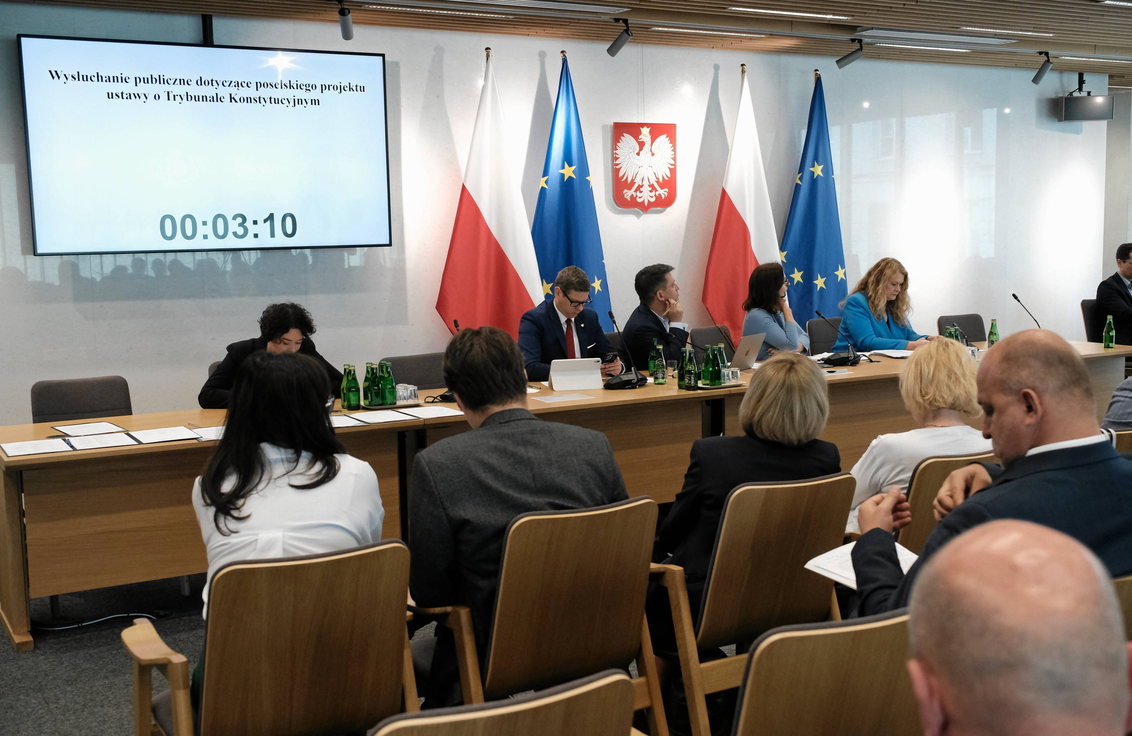 Posłowie za długim stołem na posiedzeniu sejmowej komisji, za nimi polskie godło, flaga oraz flaga Unii Europejskiej
