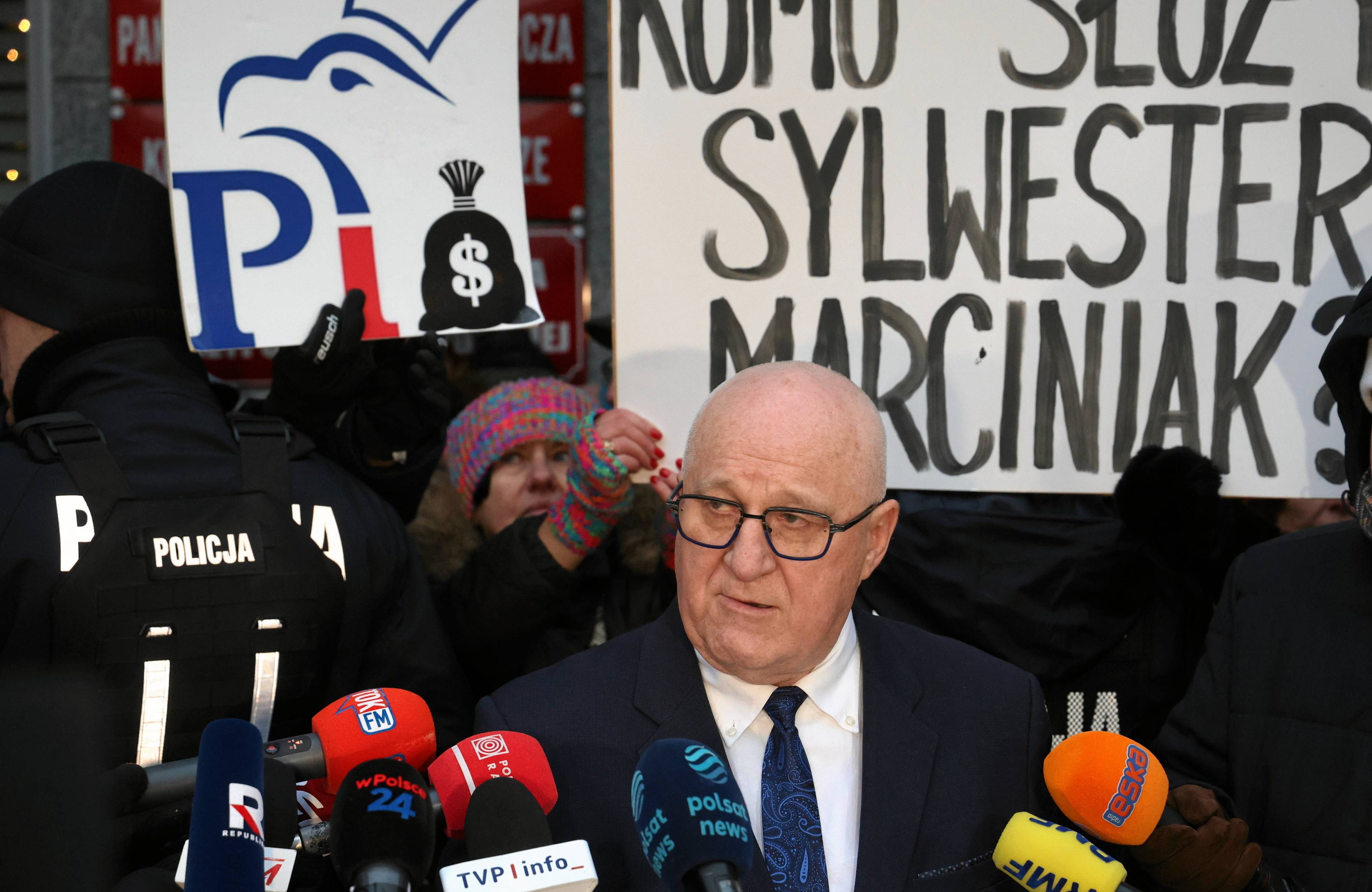 Starszy mężczyzna w okularach na konferencji prasowej, za nim logo PiS i transparent „Komu służy Sylwester Marciniak"