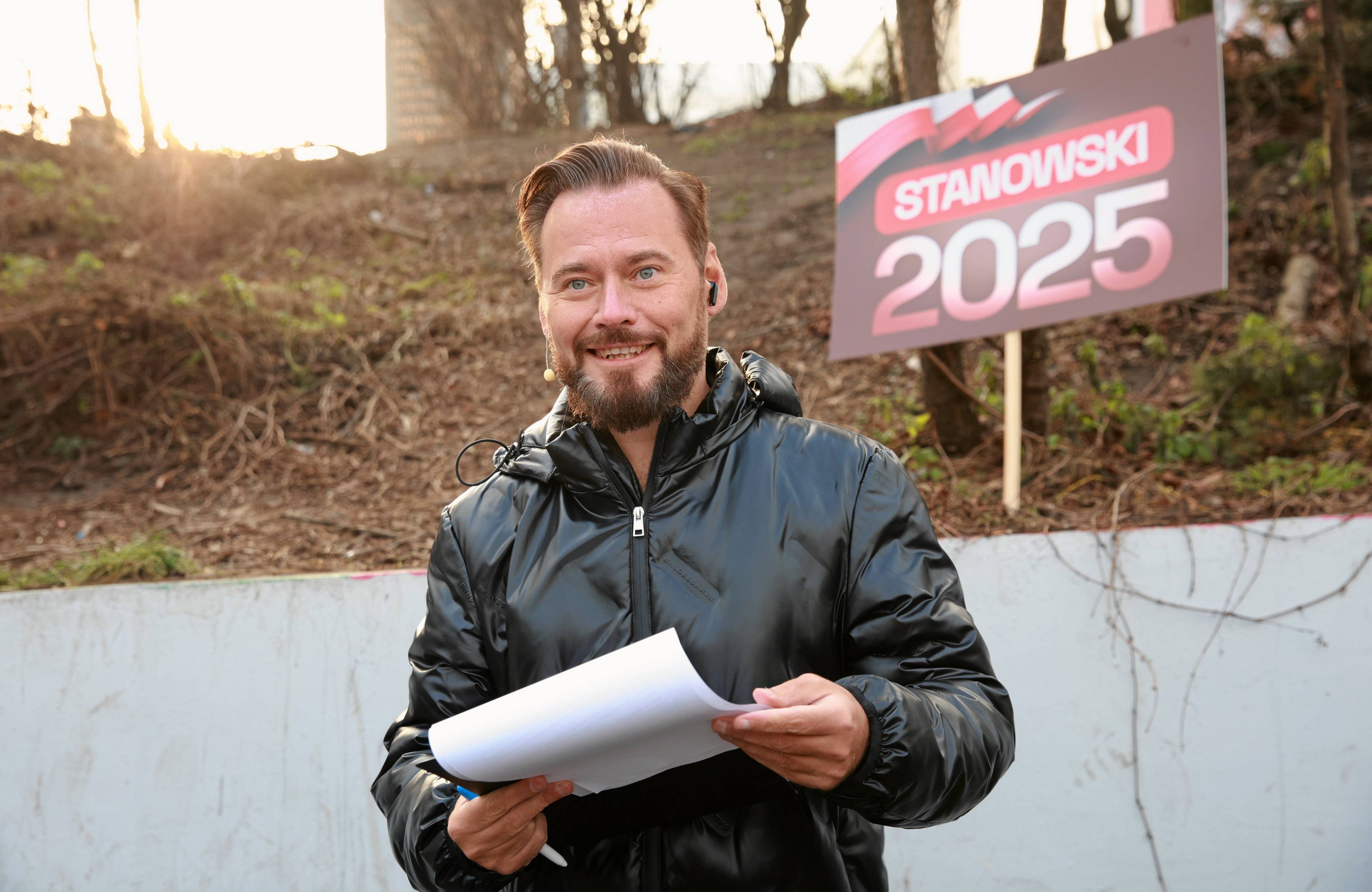 Uśmiechnięty mężczyzna z kartką i banerem z napisem „2025” za plecami. Krzysztof Stanowski