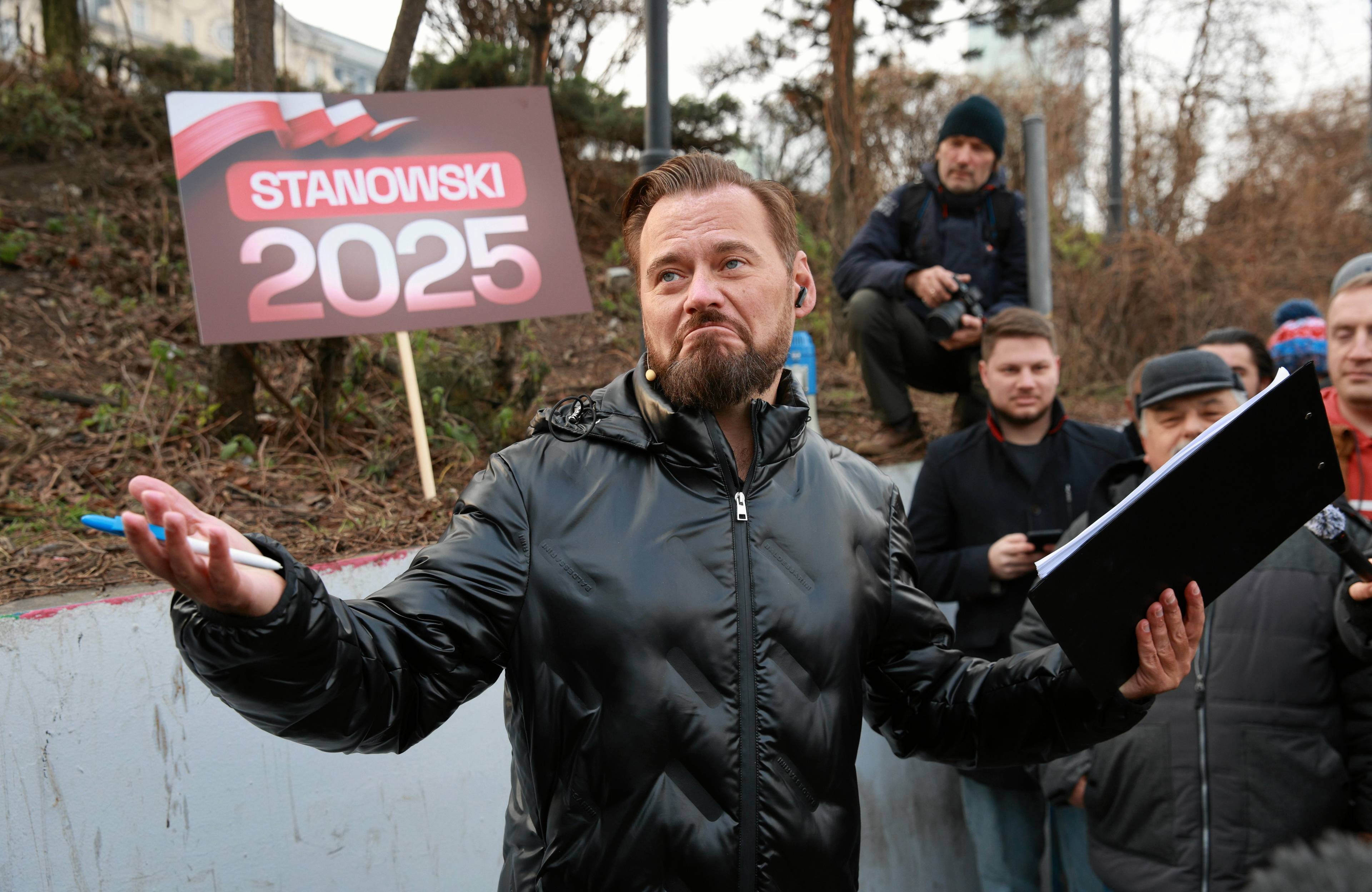 Mężczyzna w czarnej kurtce rozkłada ręce, w jednej z nich trzyma kartki. Za nim napis "Stanowski 2025"