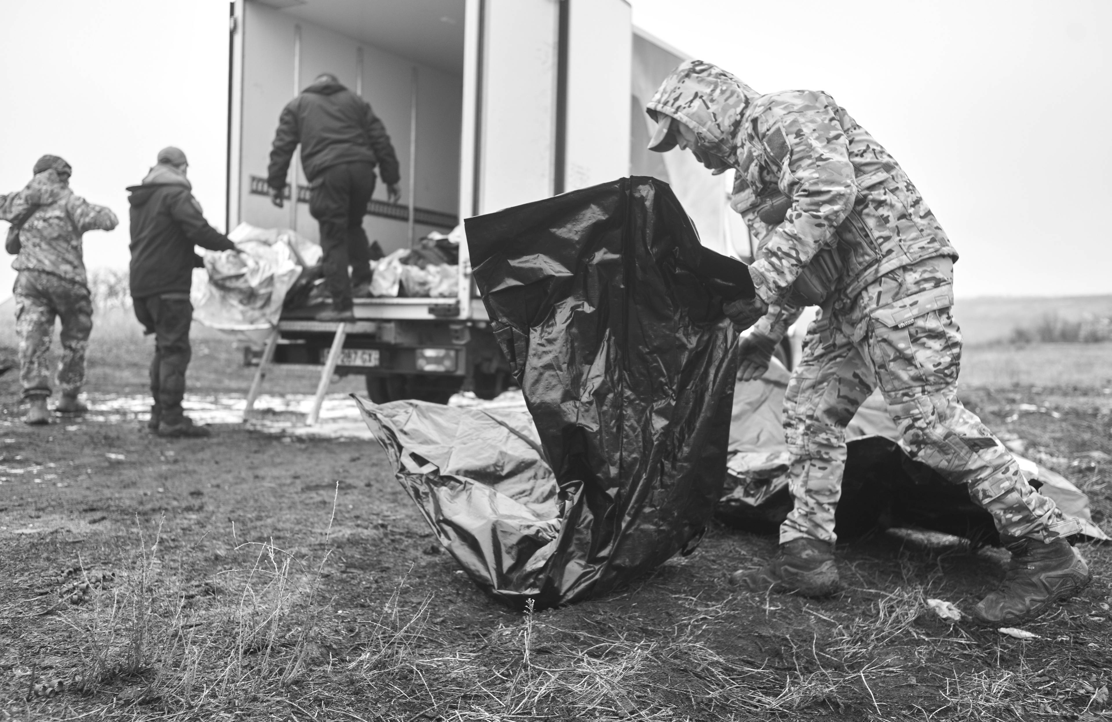 Mężczyzna w wojskowym skafandrze z kapturem niesie czarny plastikowy worek. W tle kilka postaci przy furgonetce z otwartą paką