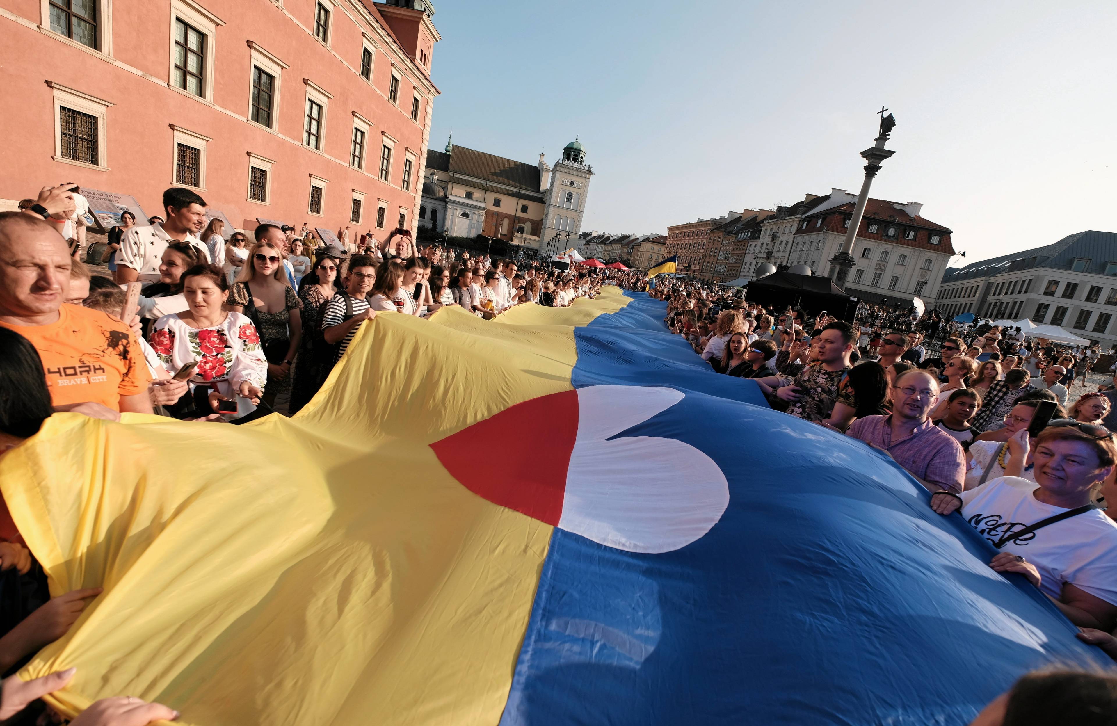 Na Placu Zamkowym w Warszawie tłum rozwija ukraińską flagę. Na środku jest serce w barwach polskiej flagi