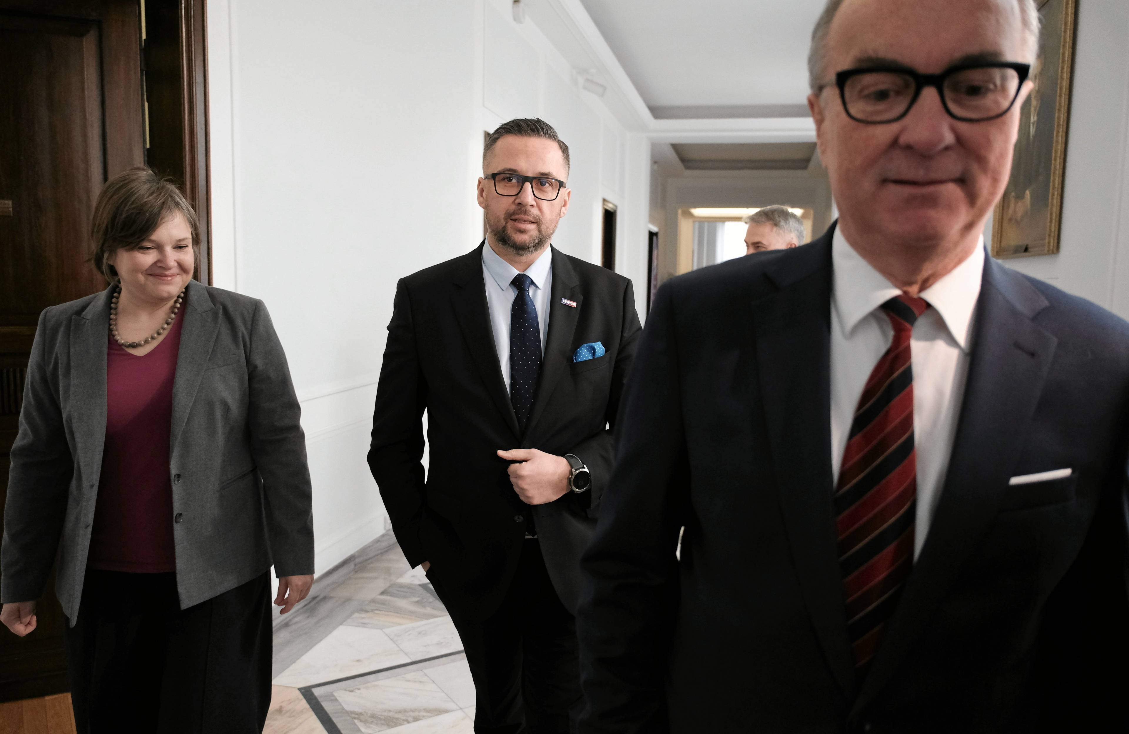 Dwaj mężczyźni w garniturach i kobieta idą korytarzem