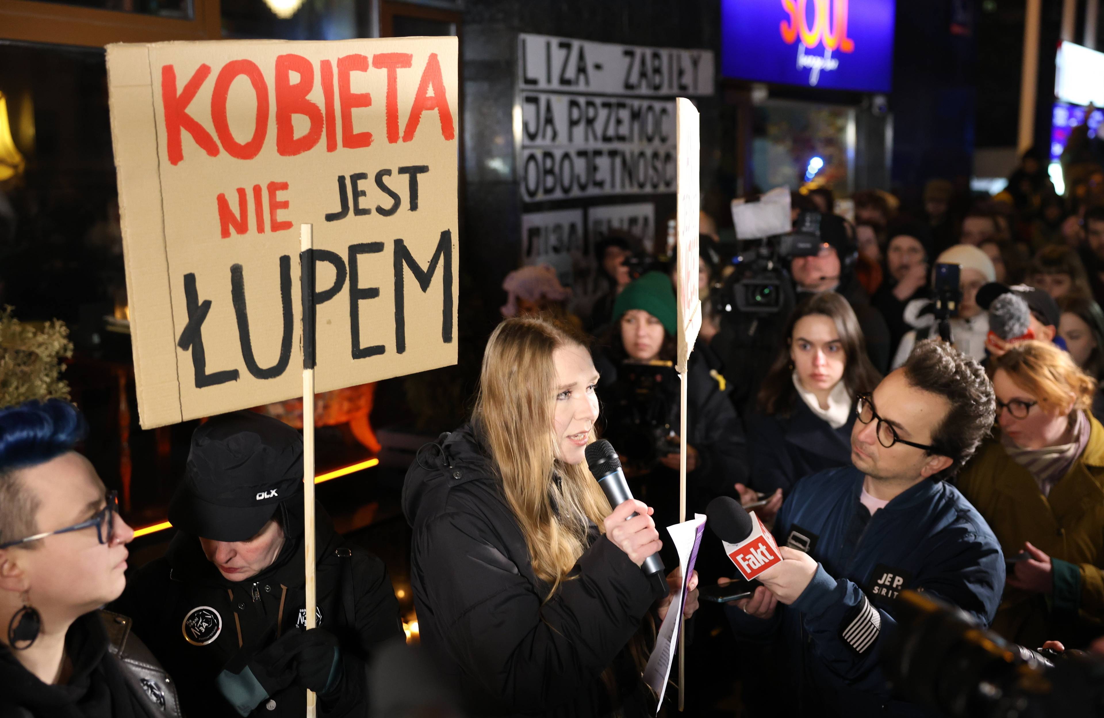 Młoda kobieta (aktywistka Maja Staśko) przemawia do mikrofonu stojąc w tłumie ludzi trzymającym transparenty. Na jednym z nich napis "Kobieta nie jest łupem"