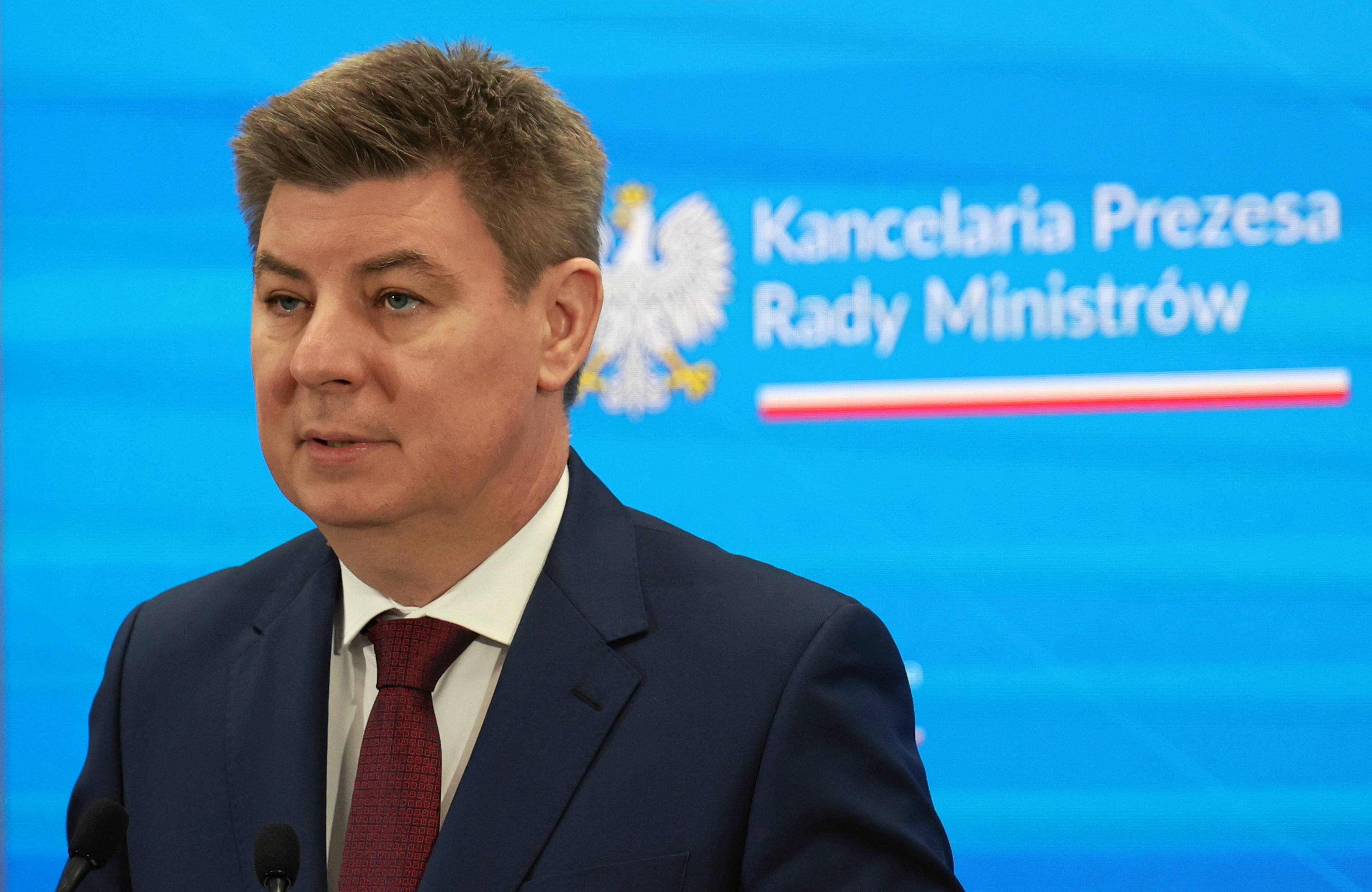 Minister Jan Grabiec. Mężczyzna w średnim wieku w garniturze i białek koszuli, na tle ścianki z napisem Kancelaria Prezesa Rady Ministrów