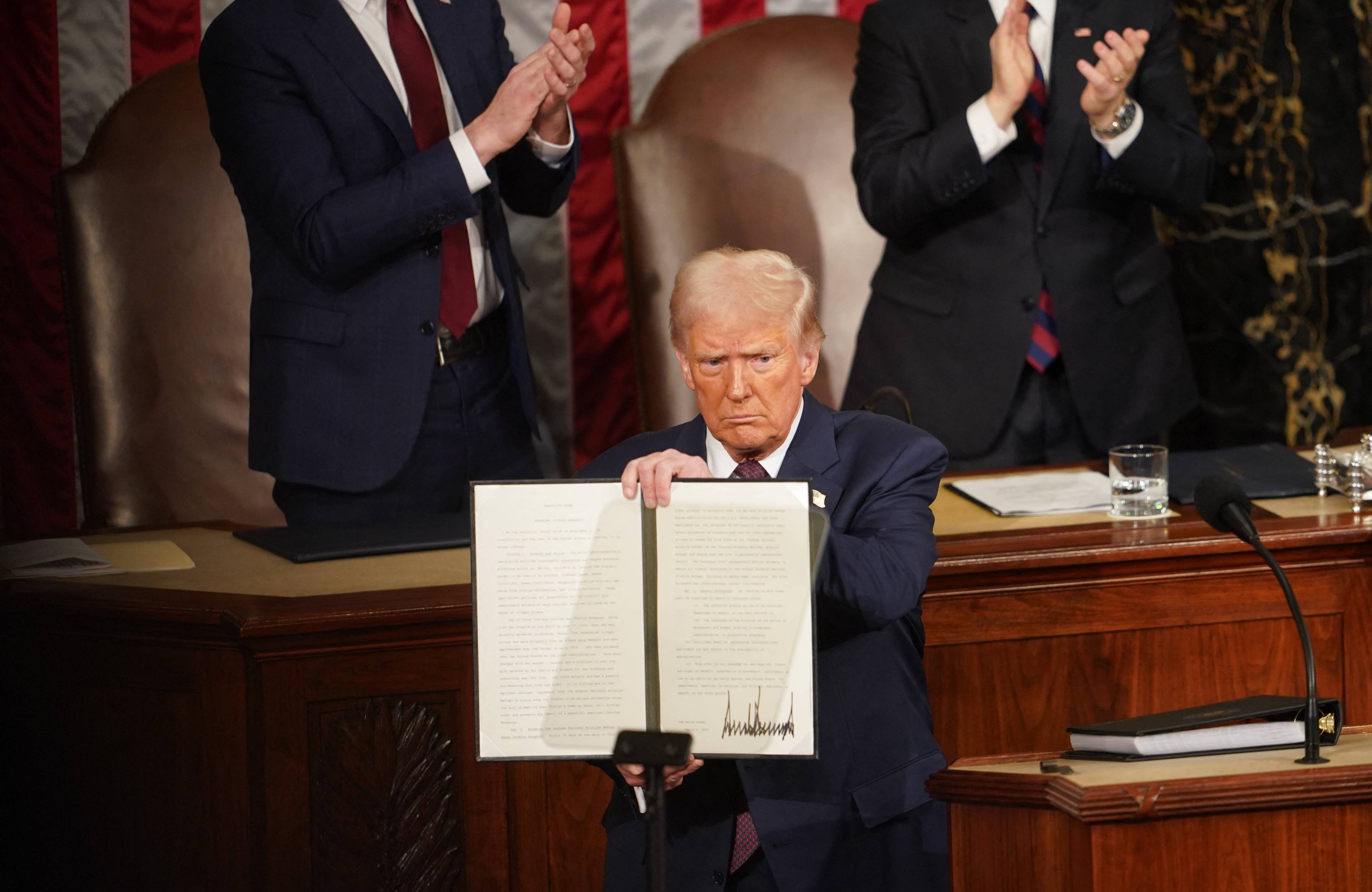 Donald Trump stoi na trybunie i pokazuje tekst rozporządzenia ze swoim podpisem