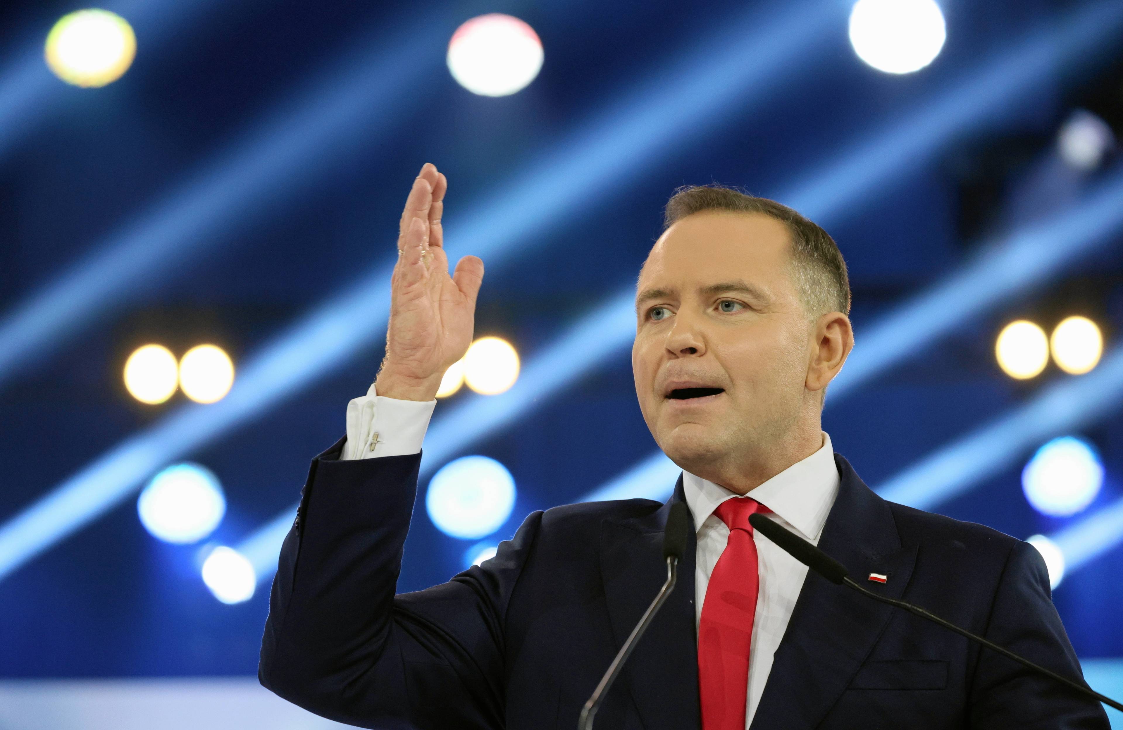 Mężczyzna z uniesioną dłonią, w garniturze i z czerwonym krawatem. Karol Nawrocki, konwencja programowa