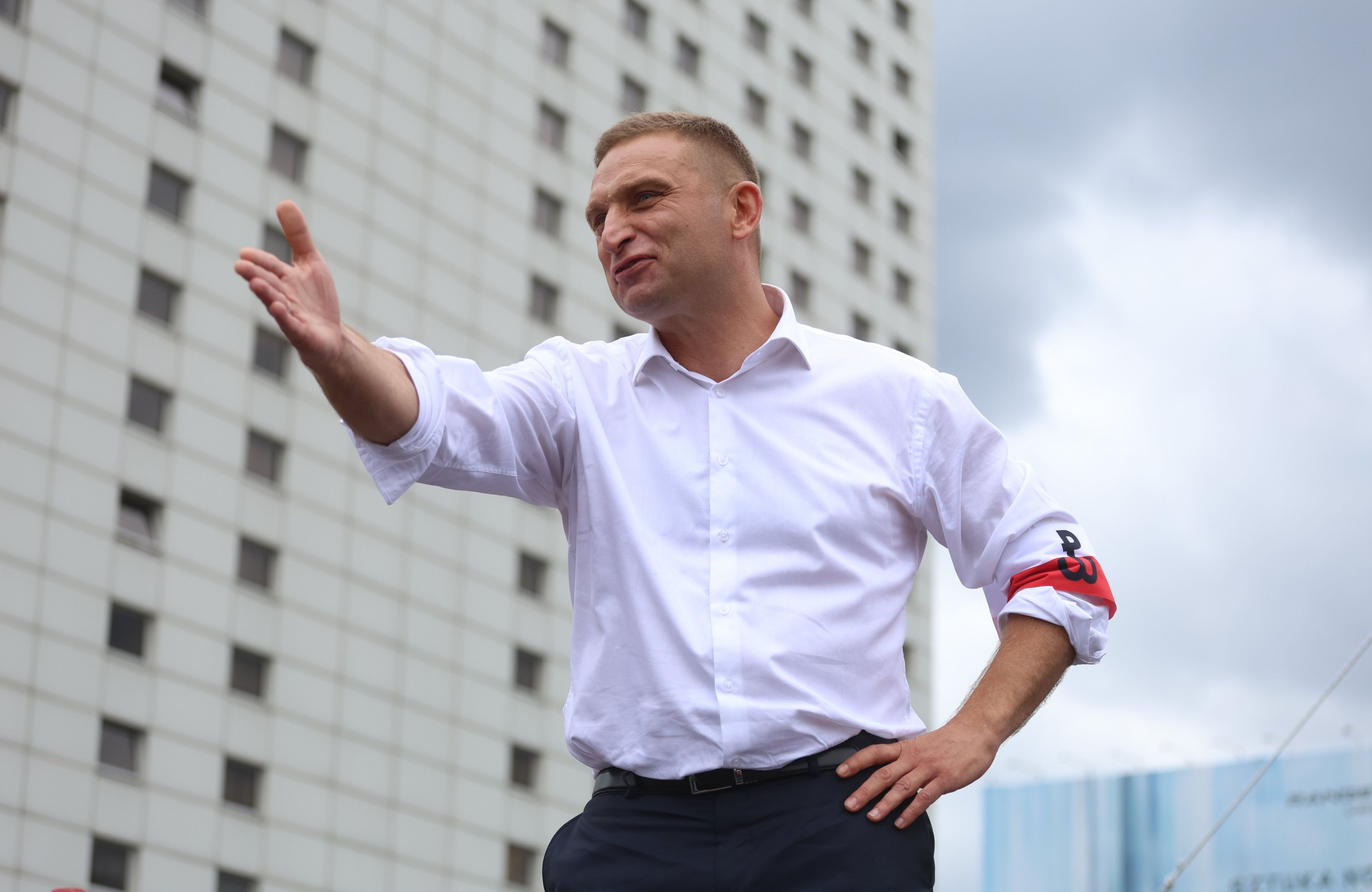 Robert Bąkiewicz w białej koszuli z opaską z logiem Polski Walczącej jedną ręką podpiera się w bok, drugą pokazuje do przodu