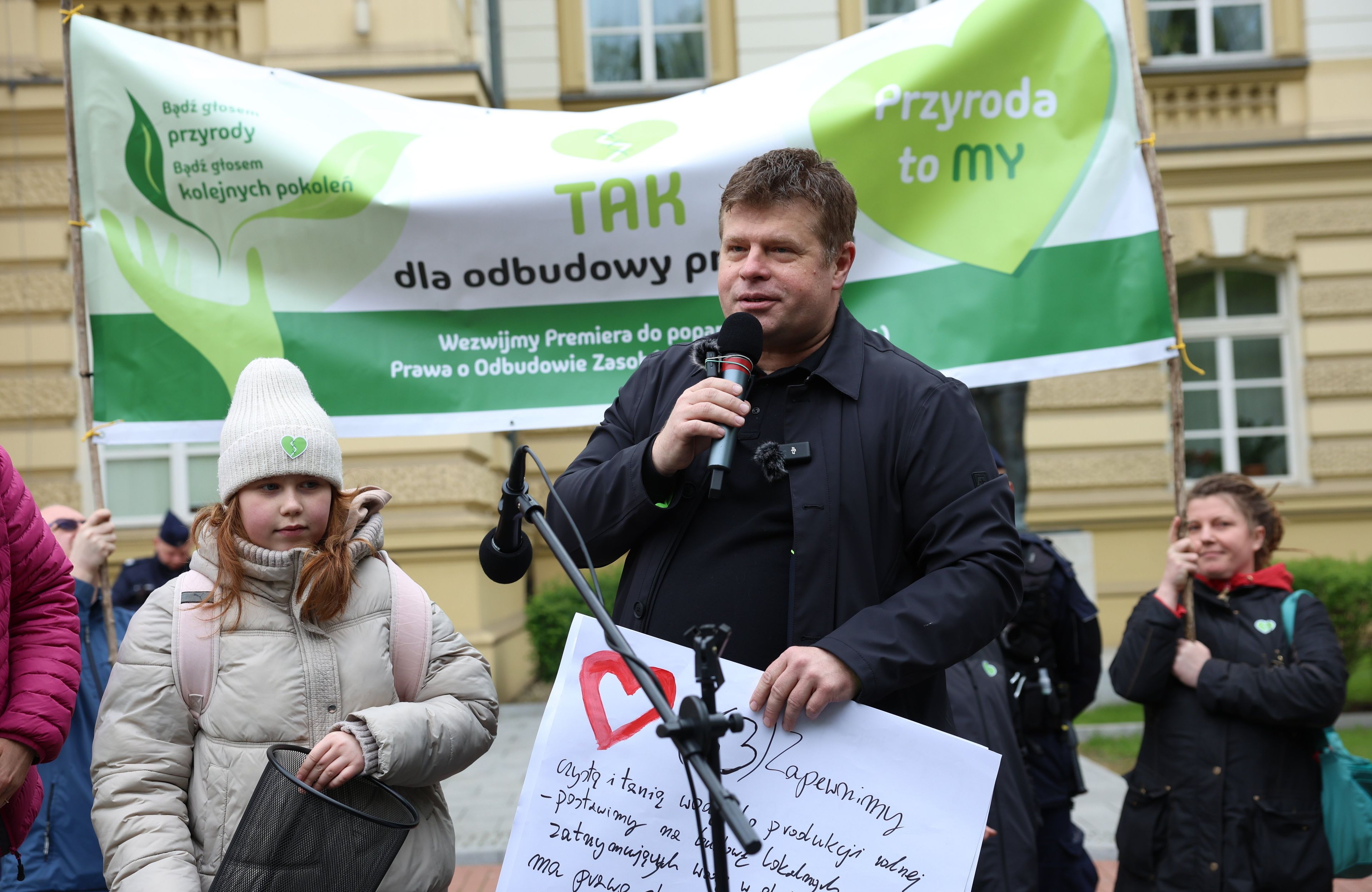 Mężczyzna przemawia na demonstracji, za nim zielone transparenty