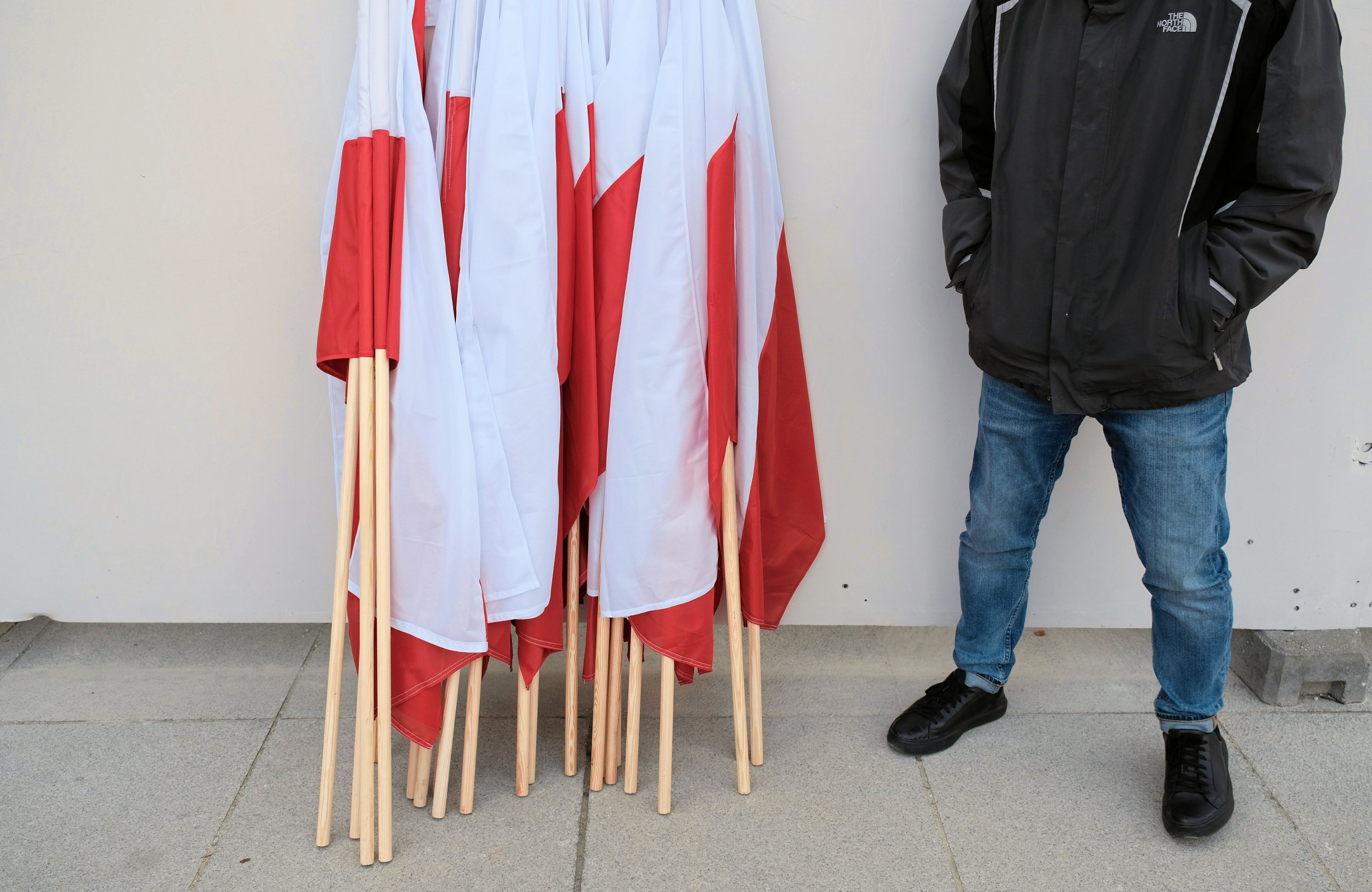 Polskie flagi stoją na drzewcach na podłodze, obok nich mężczyzna w dżinsach