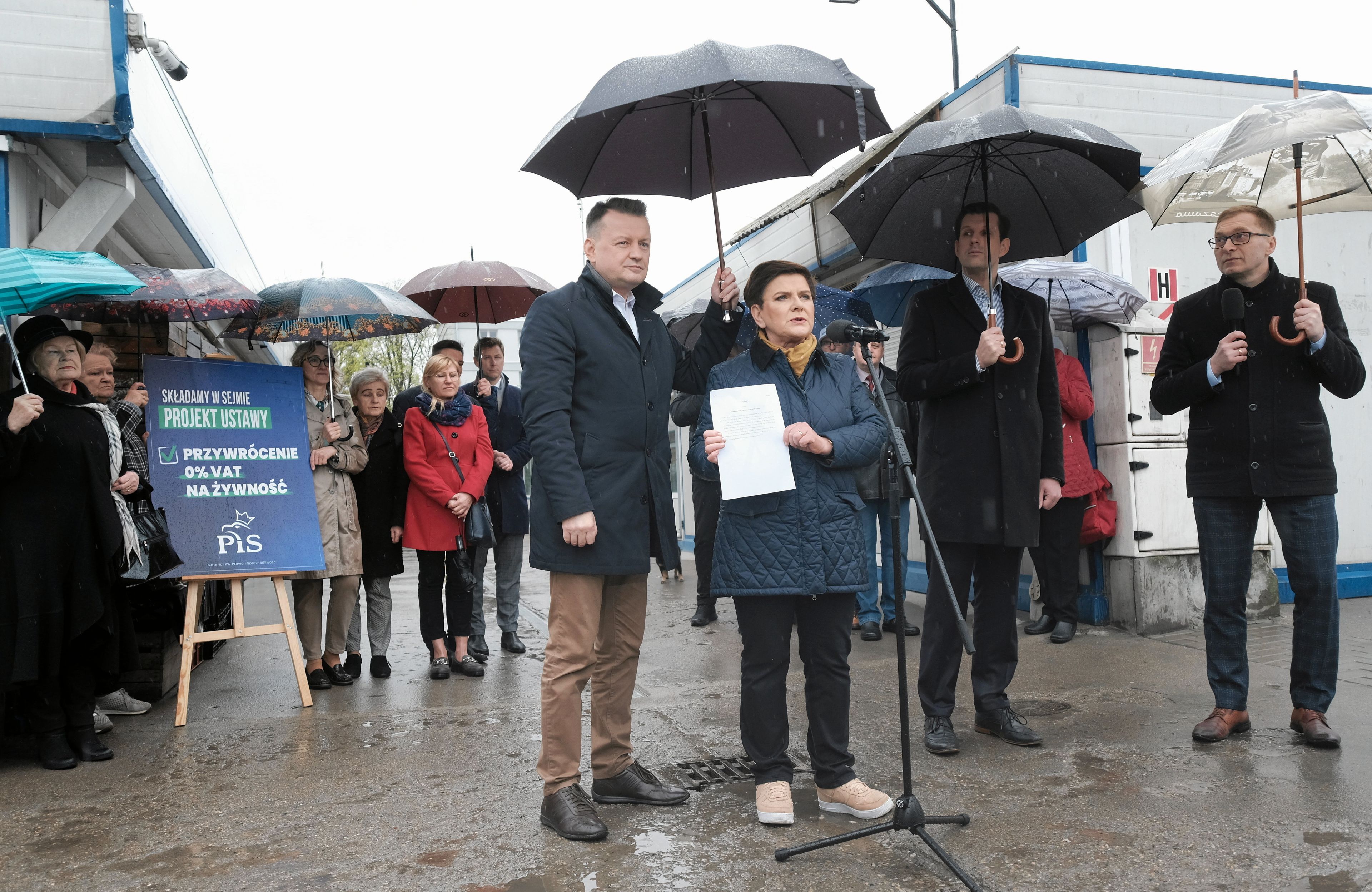 Mariusz Błaszczak i Beata Szydło pod parasolami na targowisku w Warszawie mówią o zerowym VAT na żywność.
