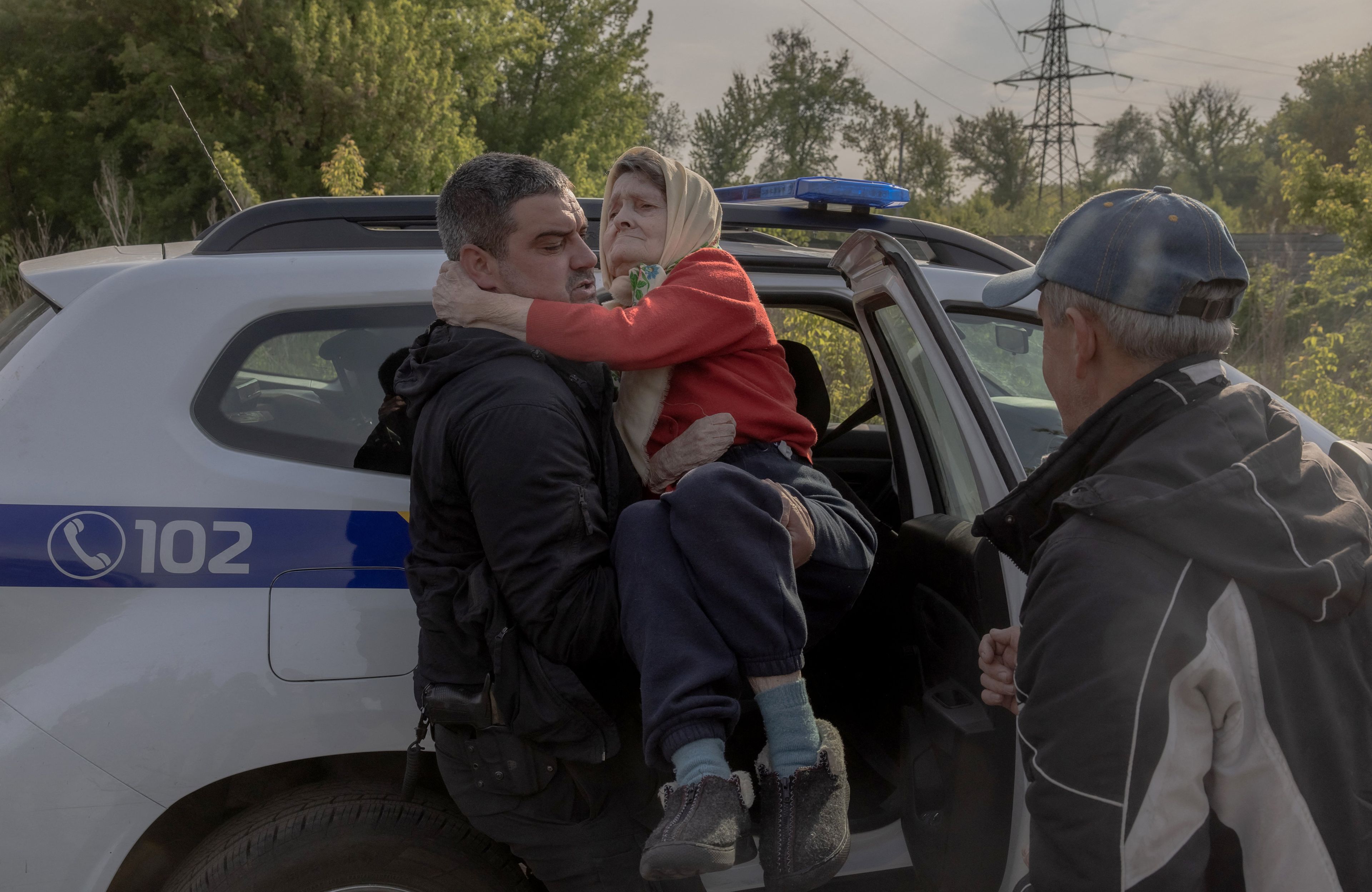 Na zdjęciu jest trzy osoby: dwóch mężczyzn i starsza pani. Jeden z mężczyzn trzyma panią na rękach, pomagał jej wyjść z samochodu policji, którym została ewakuowana