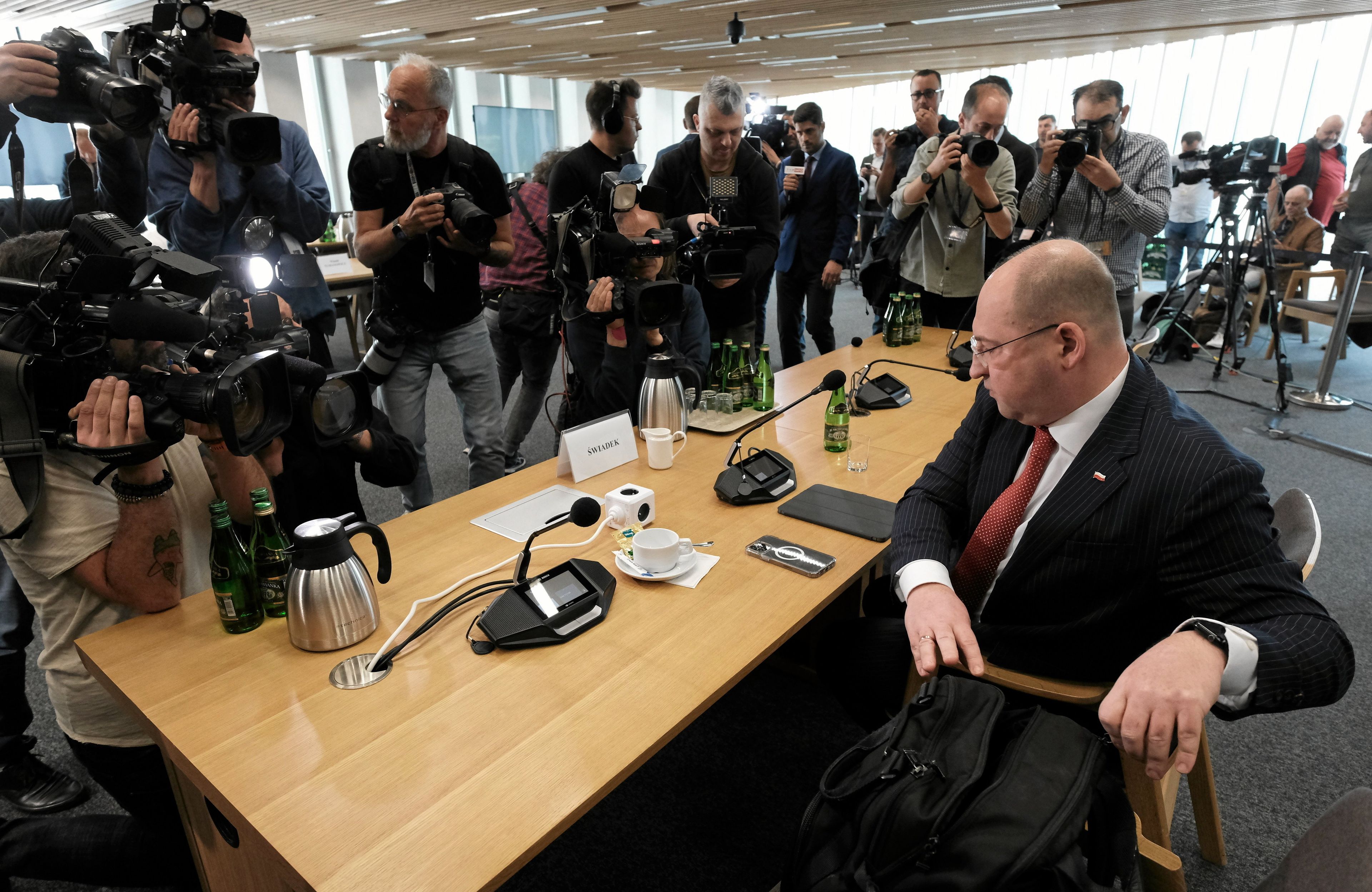 Łysiejący mężczyzna przy stole dla świadka. Przed nim tłum fotoreporterów