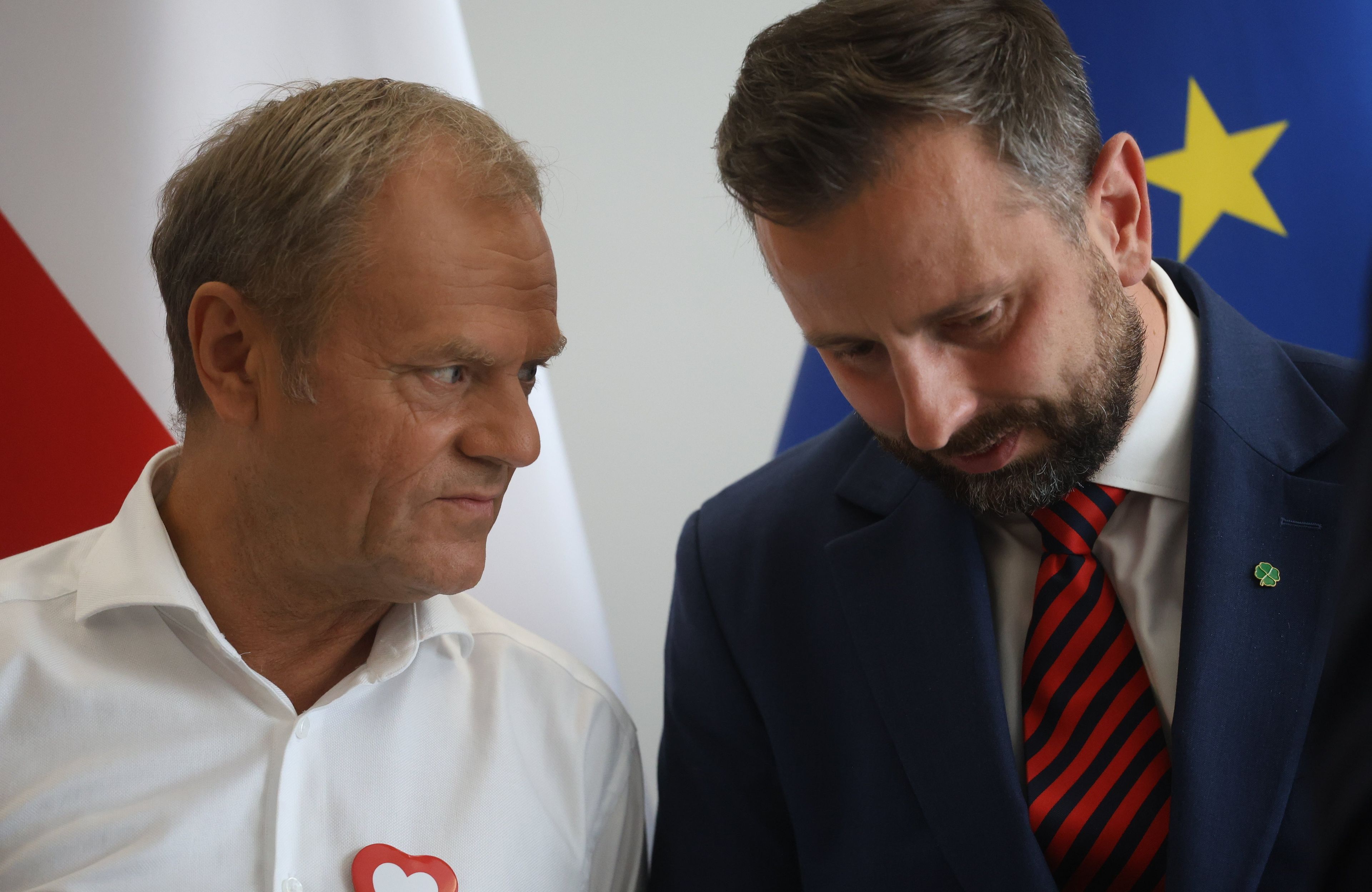 Donald Tusk na tle polskiej flagi szepcze do Władysława Kosiniaka-Kamysza (na tle flagi Unii Europejskiej), który pochyla głowę