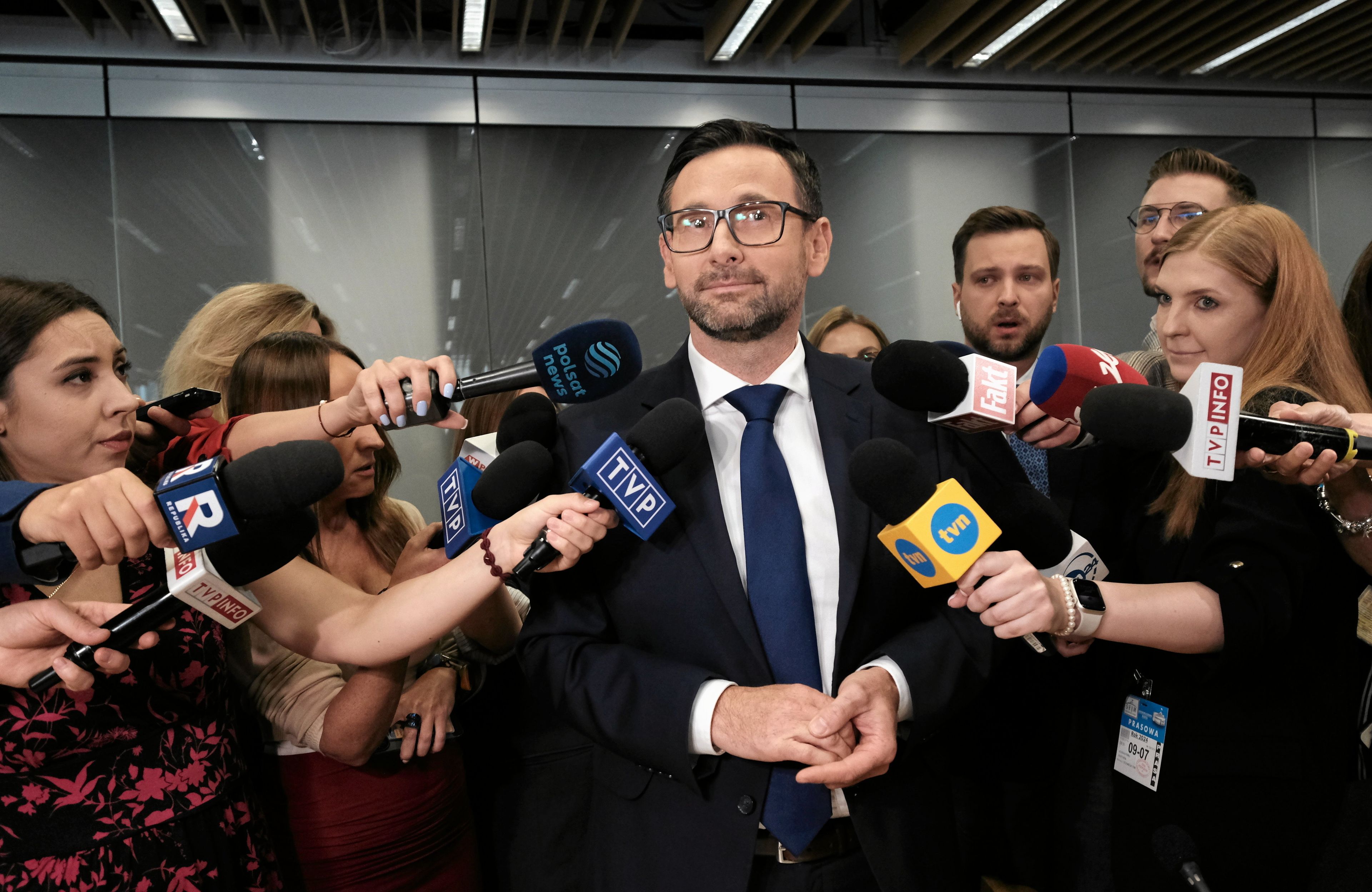 Mężczyzna w średnim wieku, ciemne włosy, okulary, garnitur, biała koszula i krawat, otoczony przez grupę dziennikarzy z mikrofonami