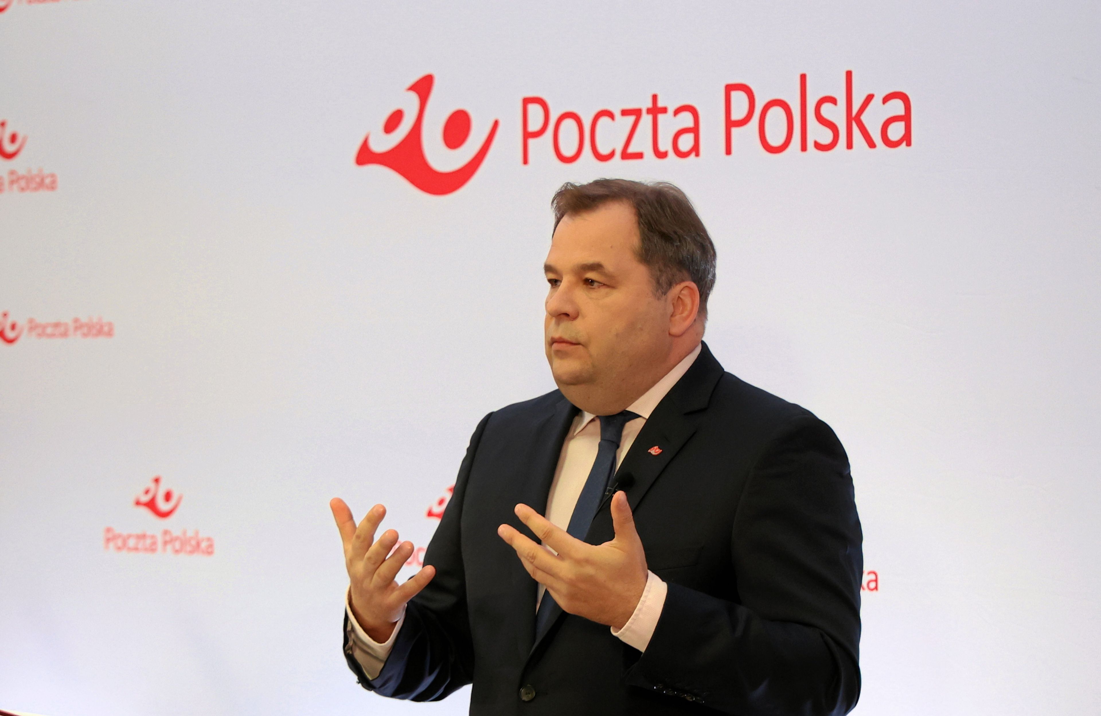 Mężczyzna w średnim wieku w ciemnym garniturze, gestykuluje na tle białej ścianki z napisem "Poczta Polska"
