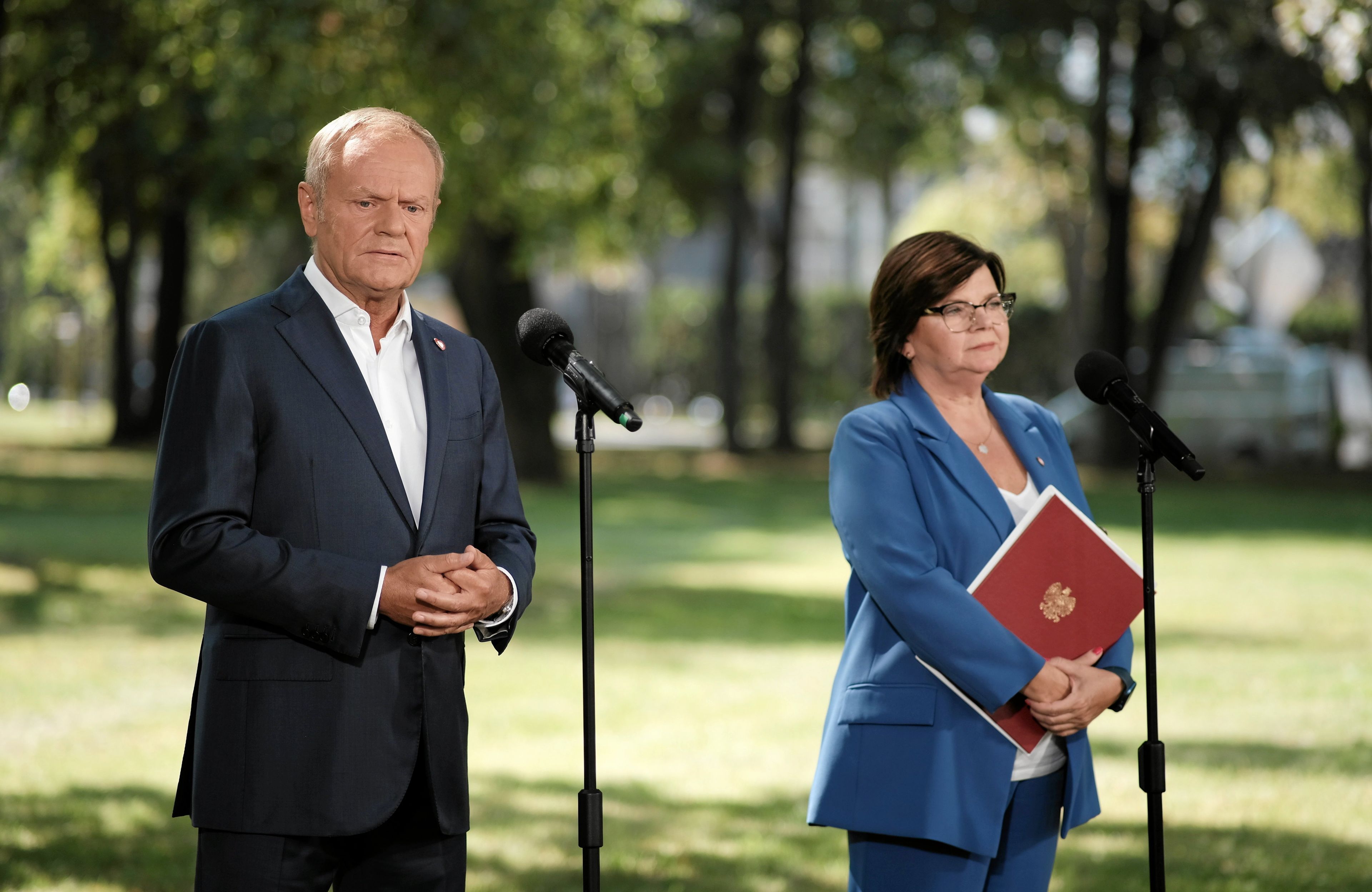 Donald Tusk i ministra zdrowia Izabela Leszczyna stoją przed mikrofonami w parku podczas konferencji prasowej. Wytyczne w sprawie aborcji
