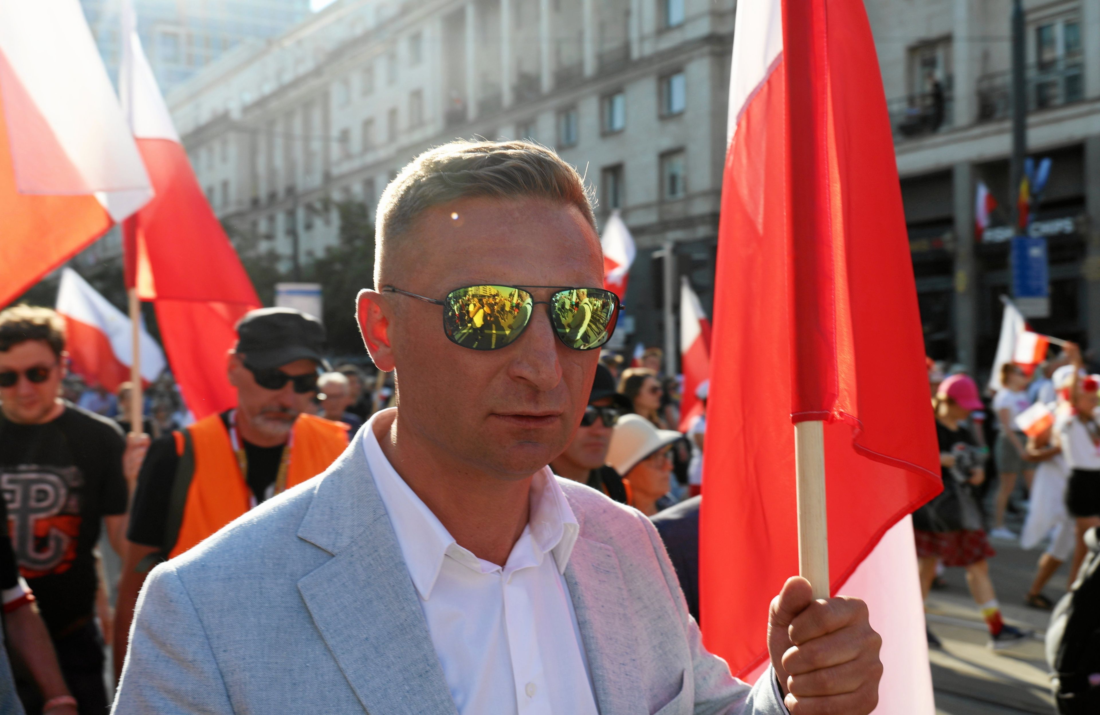 Krótko ostrzyżony mężczyzna w okularach przeciwsłonecznych i błękitnej marynarce maszeruje w tłumie ludzi z biało-czerwoną flagą w ręce