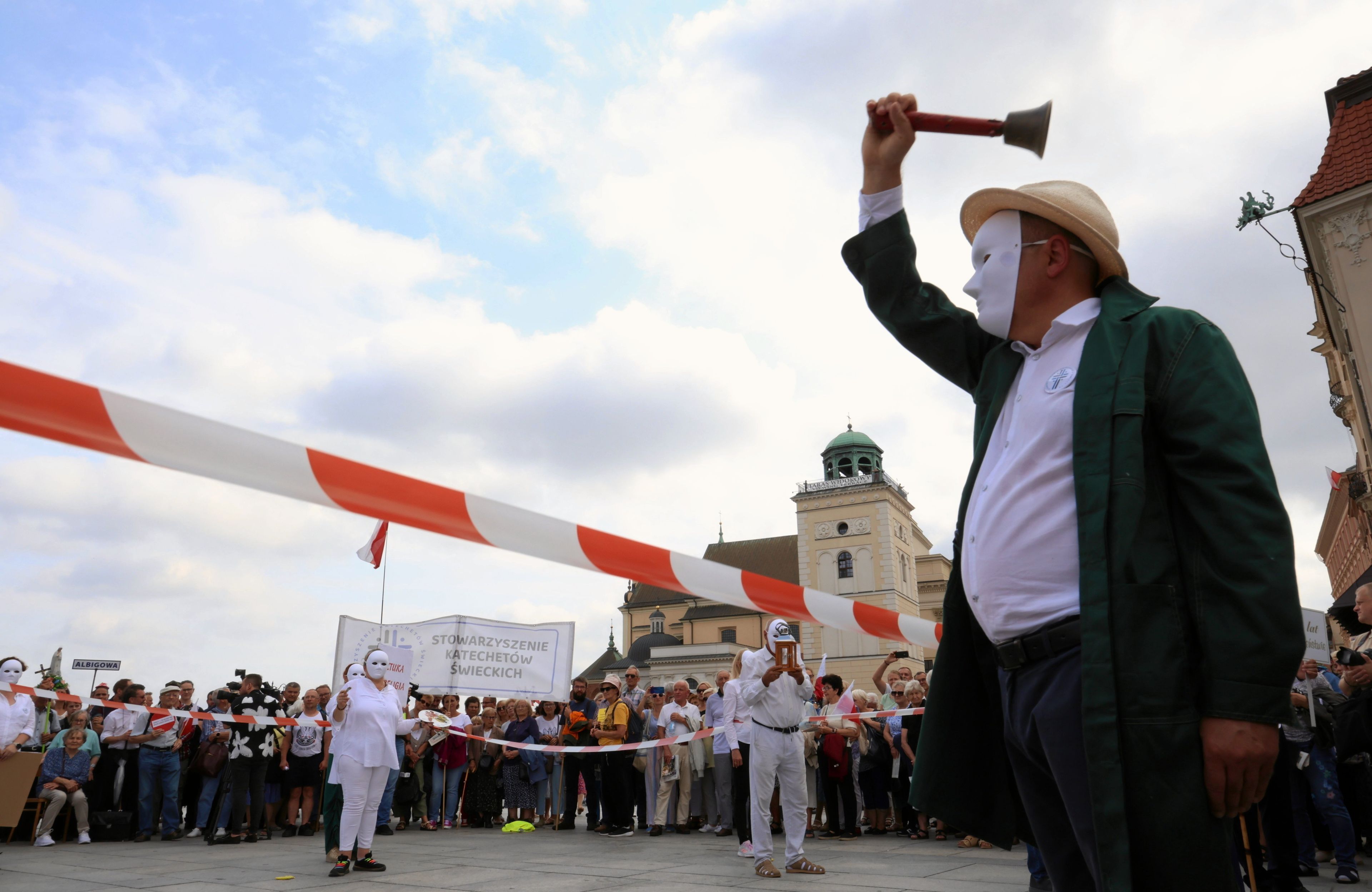 Mężczyzna w białek masce zakrywającej całą twarz macha szkolnym dzwonkiem