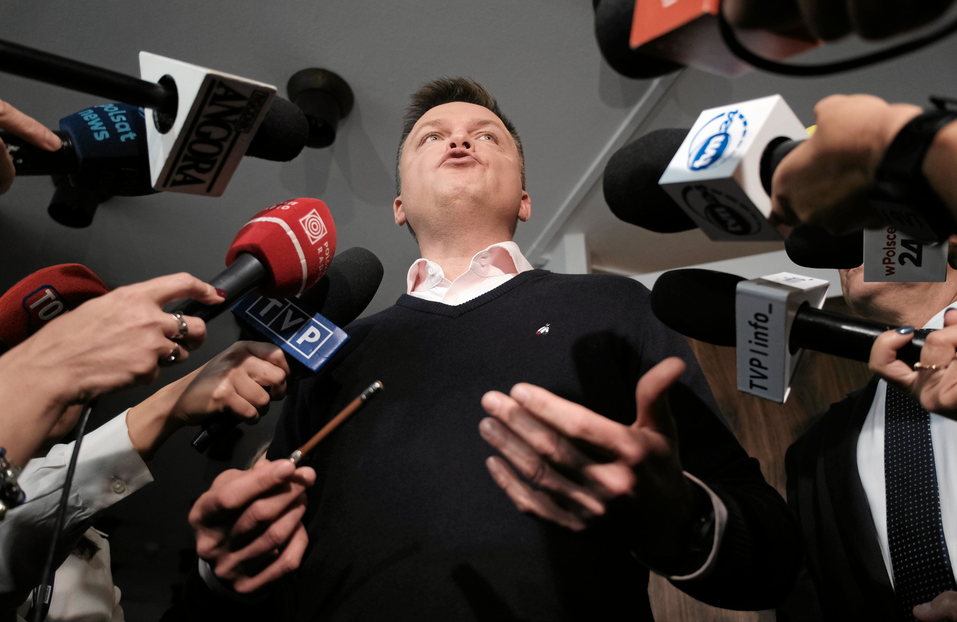 Mezczyzna w ciemnym swetrze w otoczeniu dziennikarzy