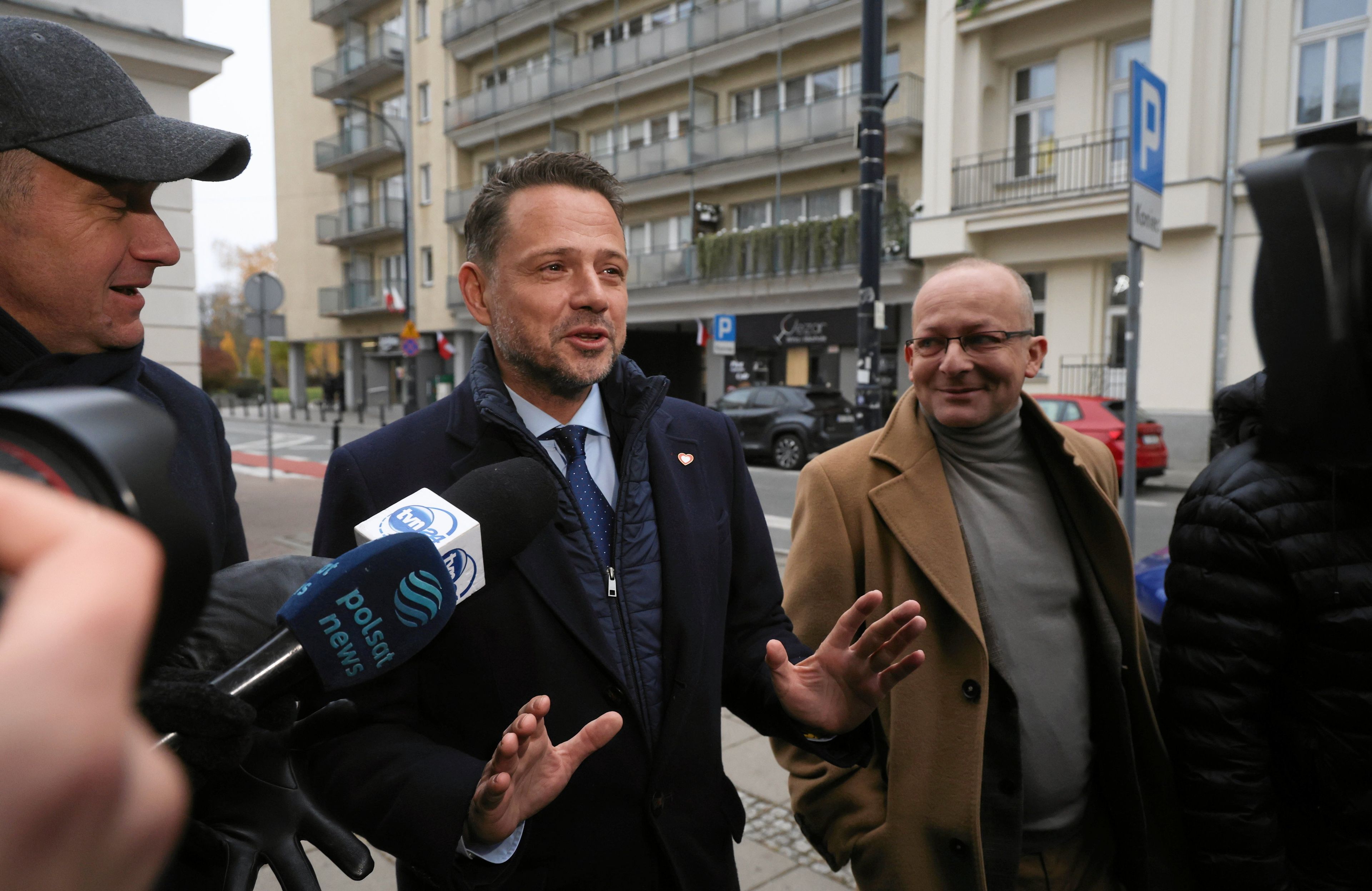 Rafał Trzaskowski z uśmiechniętą miną i rozłożonymi rękami na ulicy odpowiada na pytania mediów