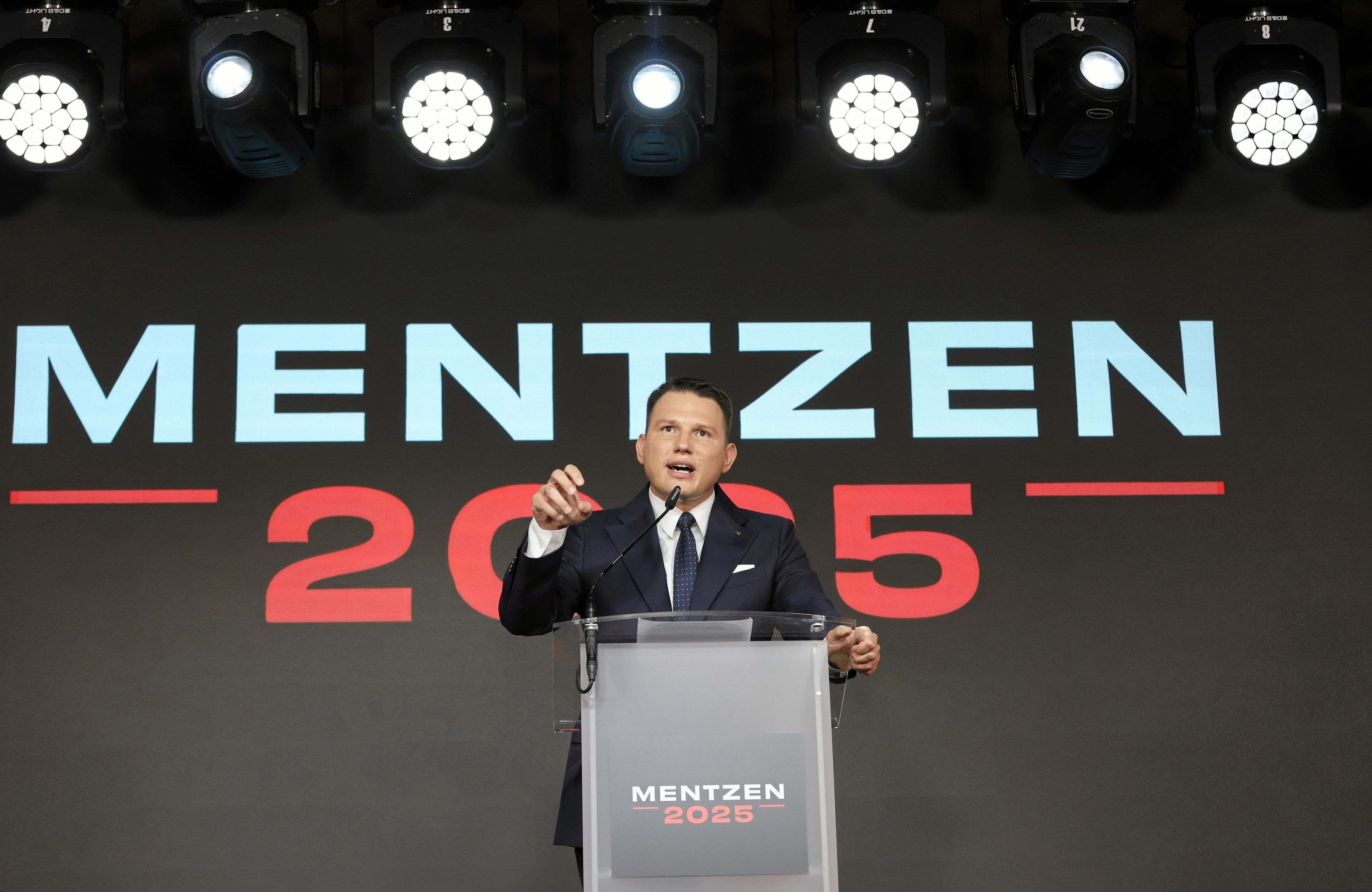 Mężczyzna w garniturze i białej koszuli stoi za mównicą i przemawia, na tle dużego napisu "Mentzen 2025"