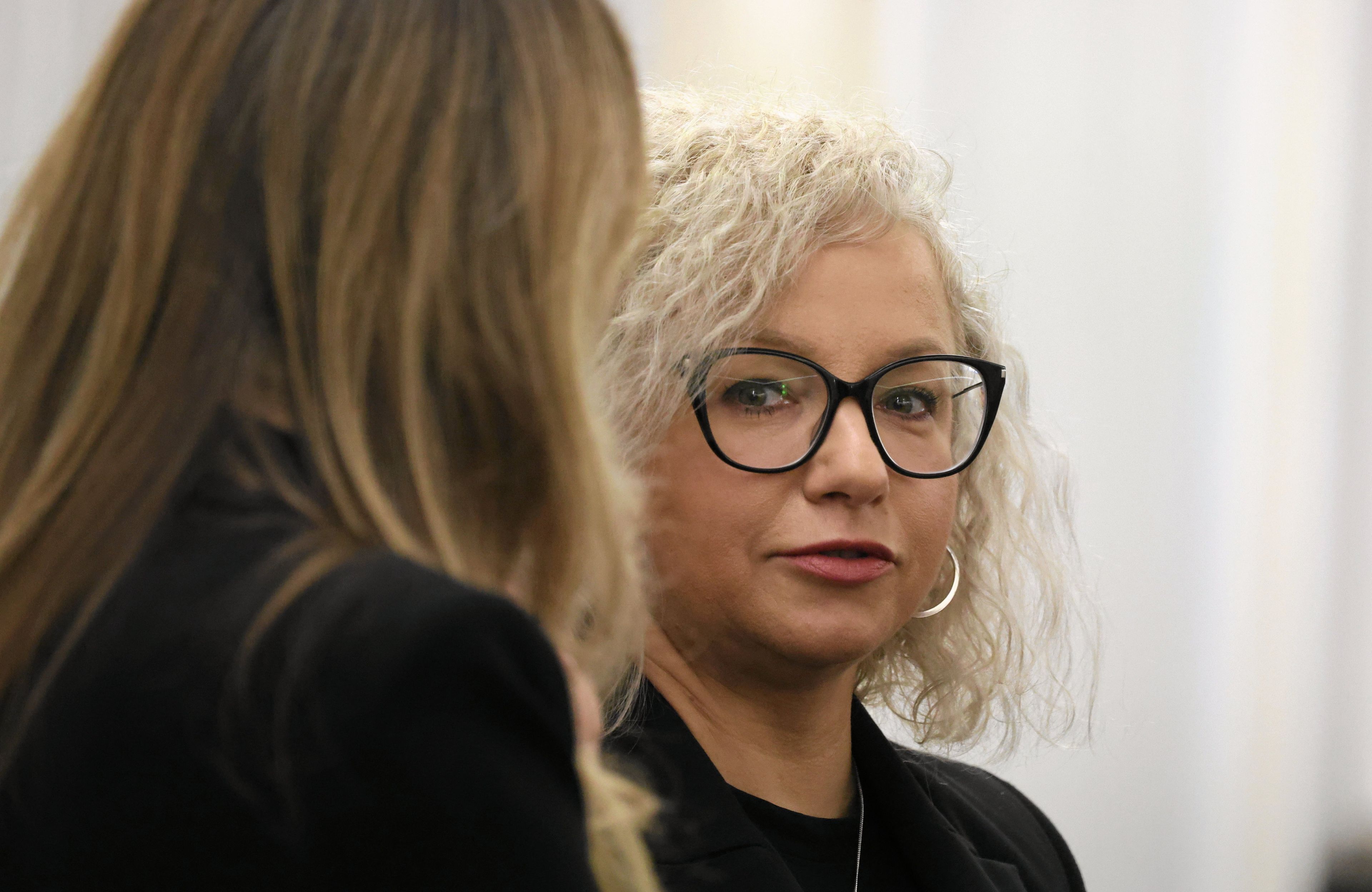 Kobieta z kręconymi blond włosami, w okularach, patrzy na kobietę z długimi włosami ujętą od tyłu.