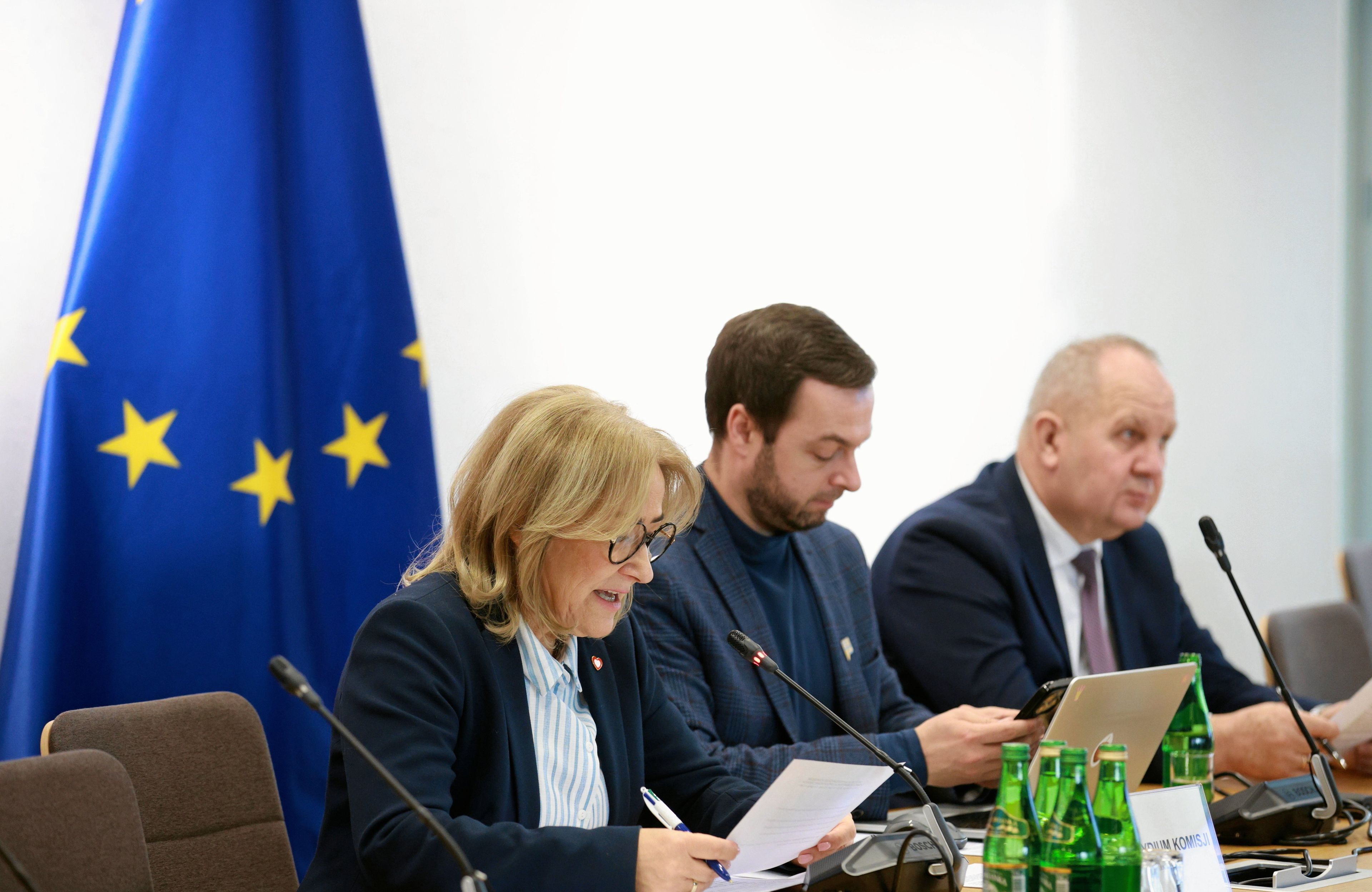Trzy osoby przy stole na posiedzeniu komisji sejmowej. Za nimi flaga UE