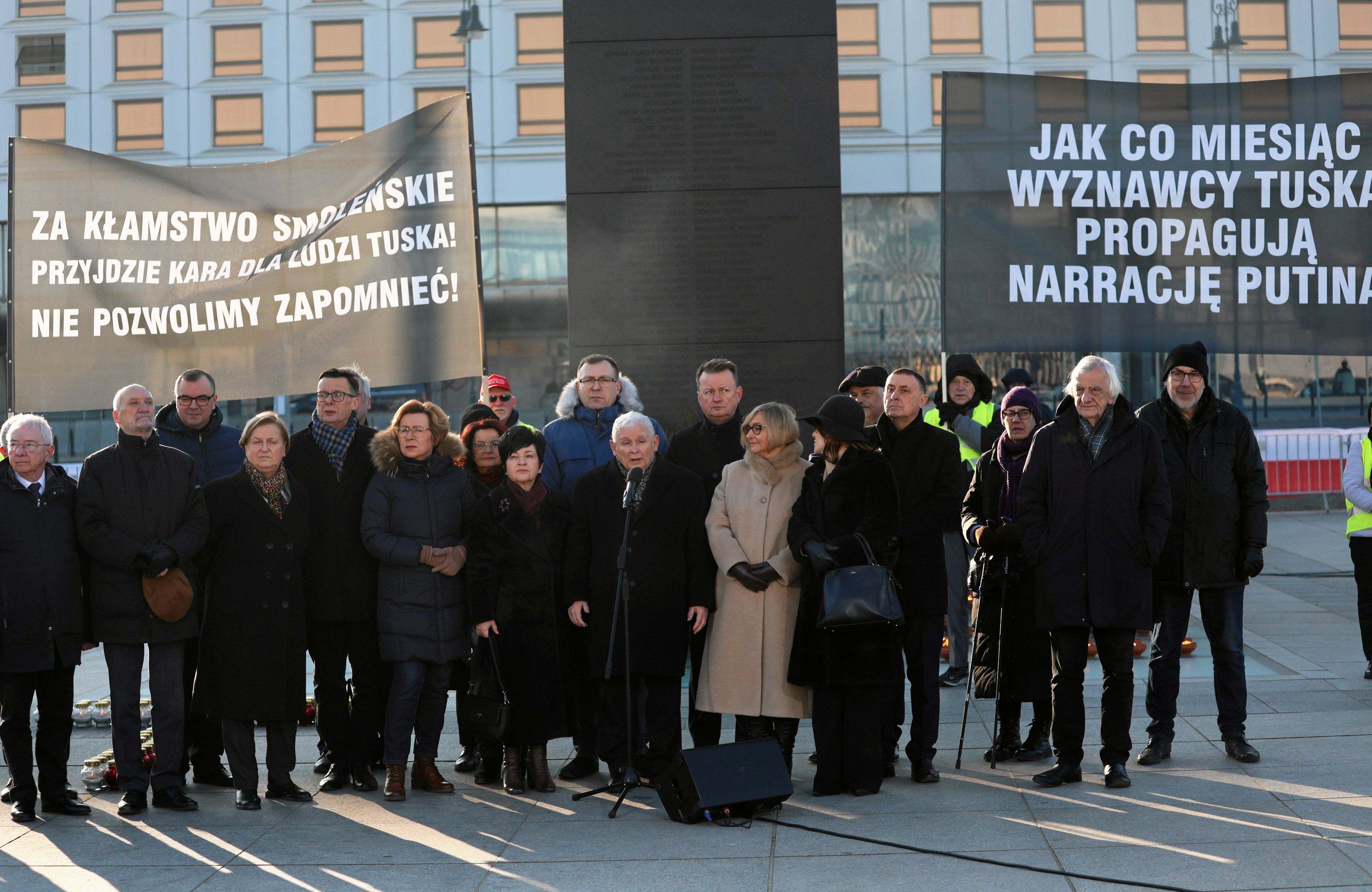 Grupa ludzi w zimowych okryciach stoi na placu, pośrodku niski mężczyzna, Jarosław Kaczyński, przemawia