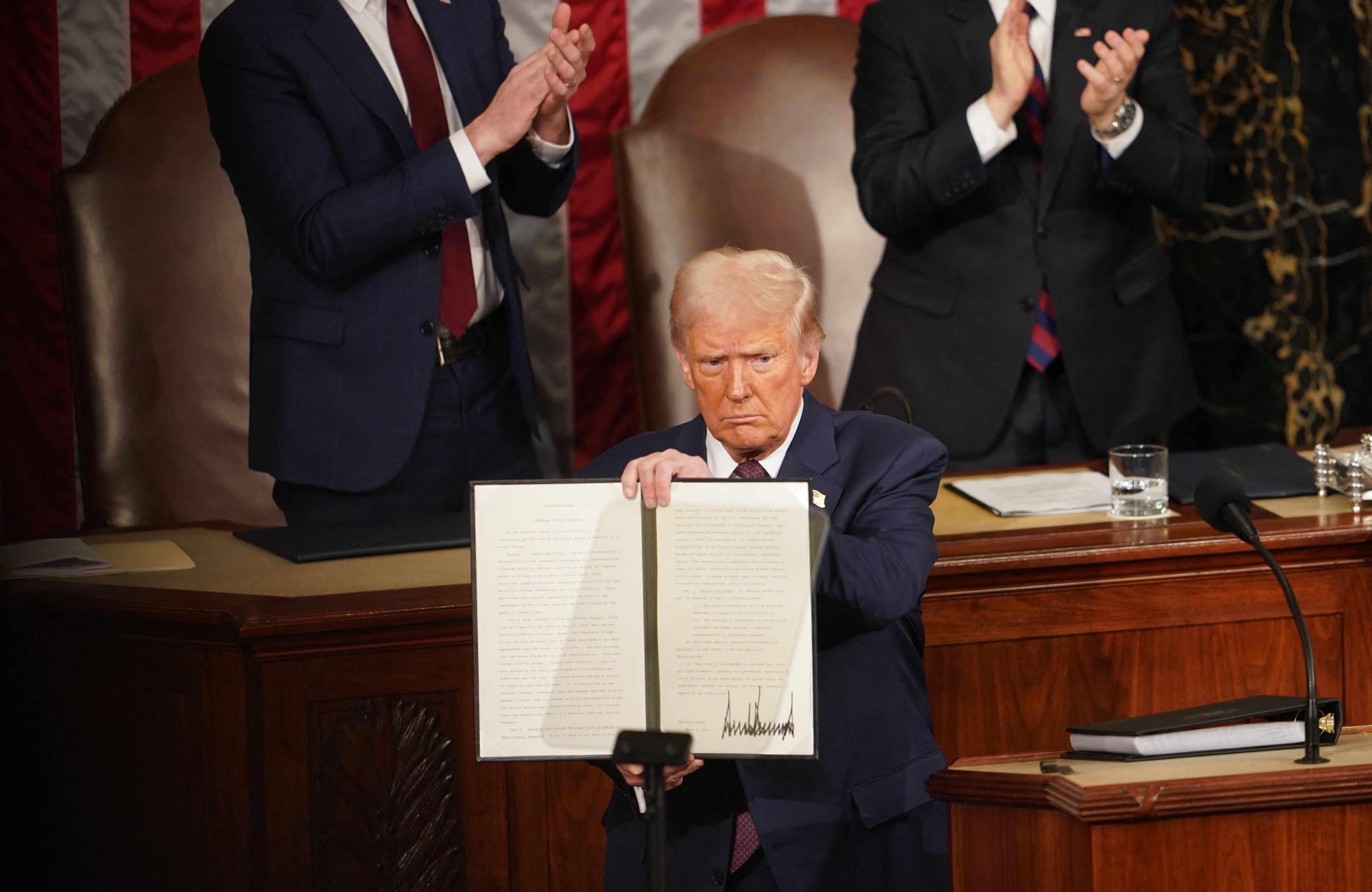 Donald Trump stoi na trybunie i pokazuje tekst rozporządzenia ze swoim podpisem