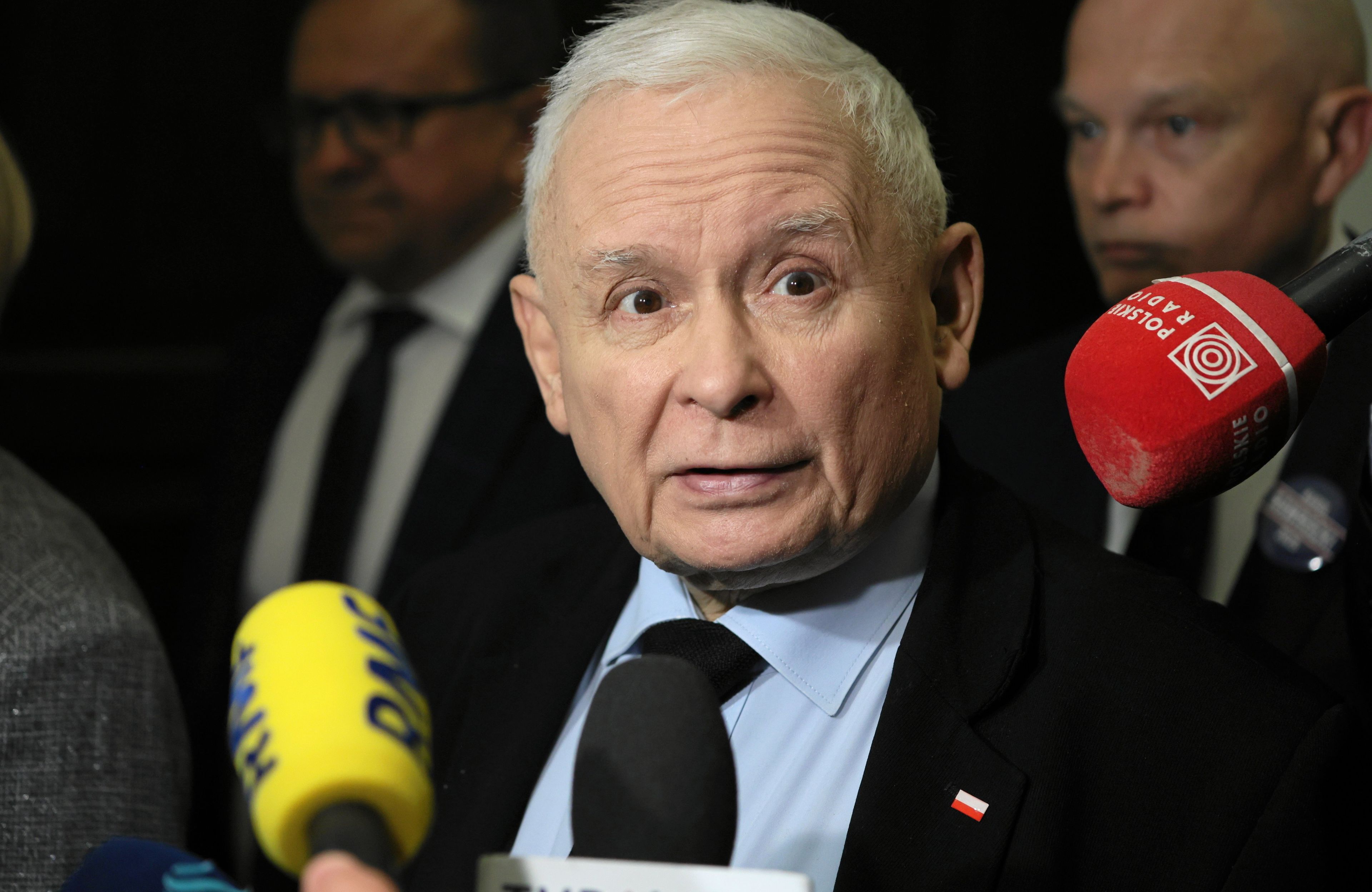 Starszy mężczyzna w garniturze i czarnym krawacie (Jarosław Kaczyński) mówi otoczony dziennikarskimi mikrofonami