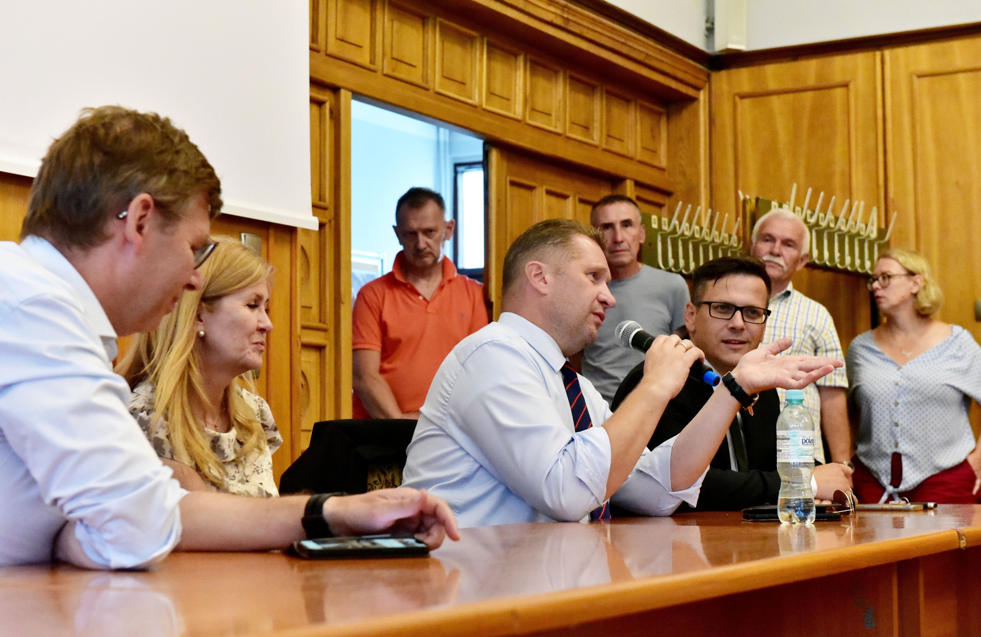 Gestykulujący mężczyzna w koszuli i krawacie trzyma mikrofon, siedzi przy stole z innymi wpatrzonymi w niego, z tyłu stoją cztery osoby