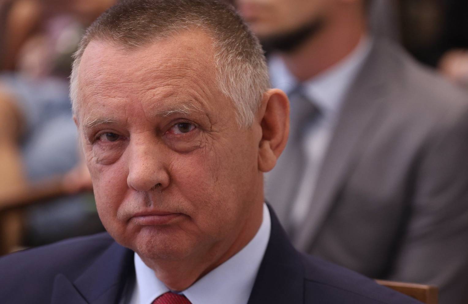 Prezes NIK Marian Banaś w garniturze i czerwonym krawacie spogląda groźnym wzrokiem w kamerę.