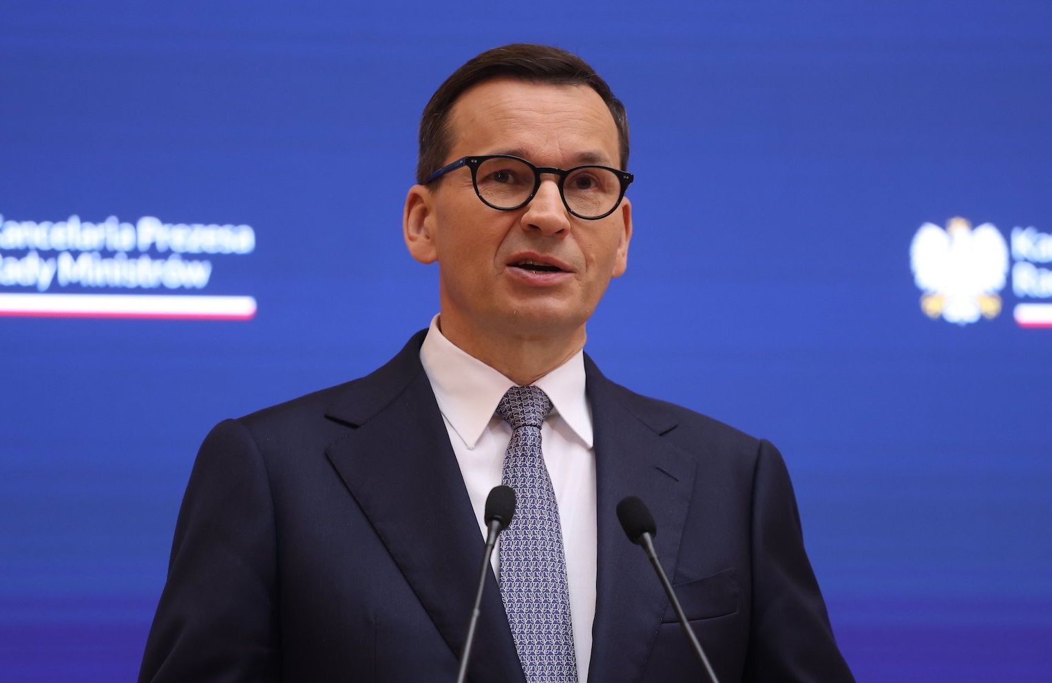 Premier Mateusz Morawiecki na konferencji prasowej w Kancelarii Premiera. Przemawia do mikrofonów na tle ścianki z logo KPRM, ubrany w granatowy garnitur, niebieski krawat i białą koszulę. Szeroko rozkłada ręce w geście bezradności i niewinności.