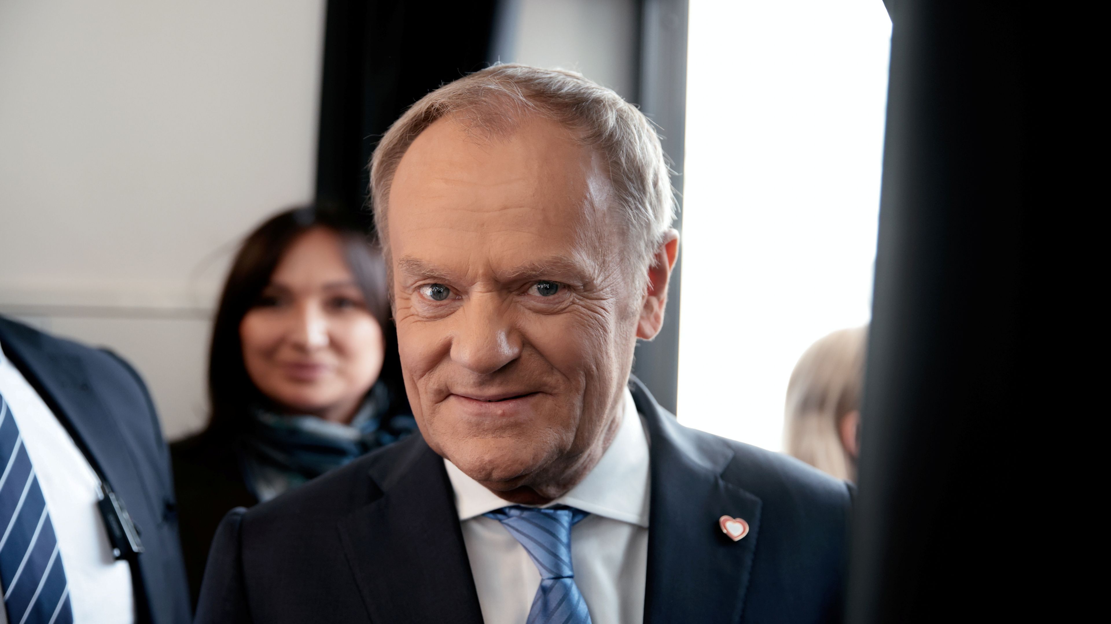 Mężczyzna w garniturze i niebieskim krawacie uśmiecha się do aparatu. W tle widać kobietę i inne osoby w pomieszczeniu oświetlonym naturalnym światłem wpadającym przez okno.