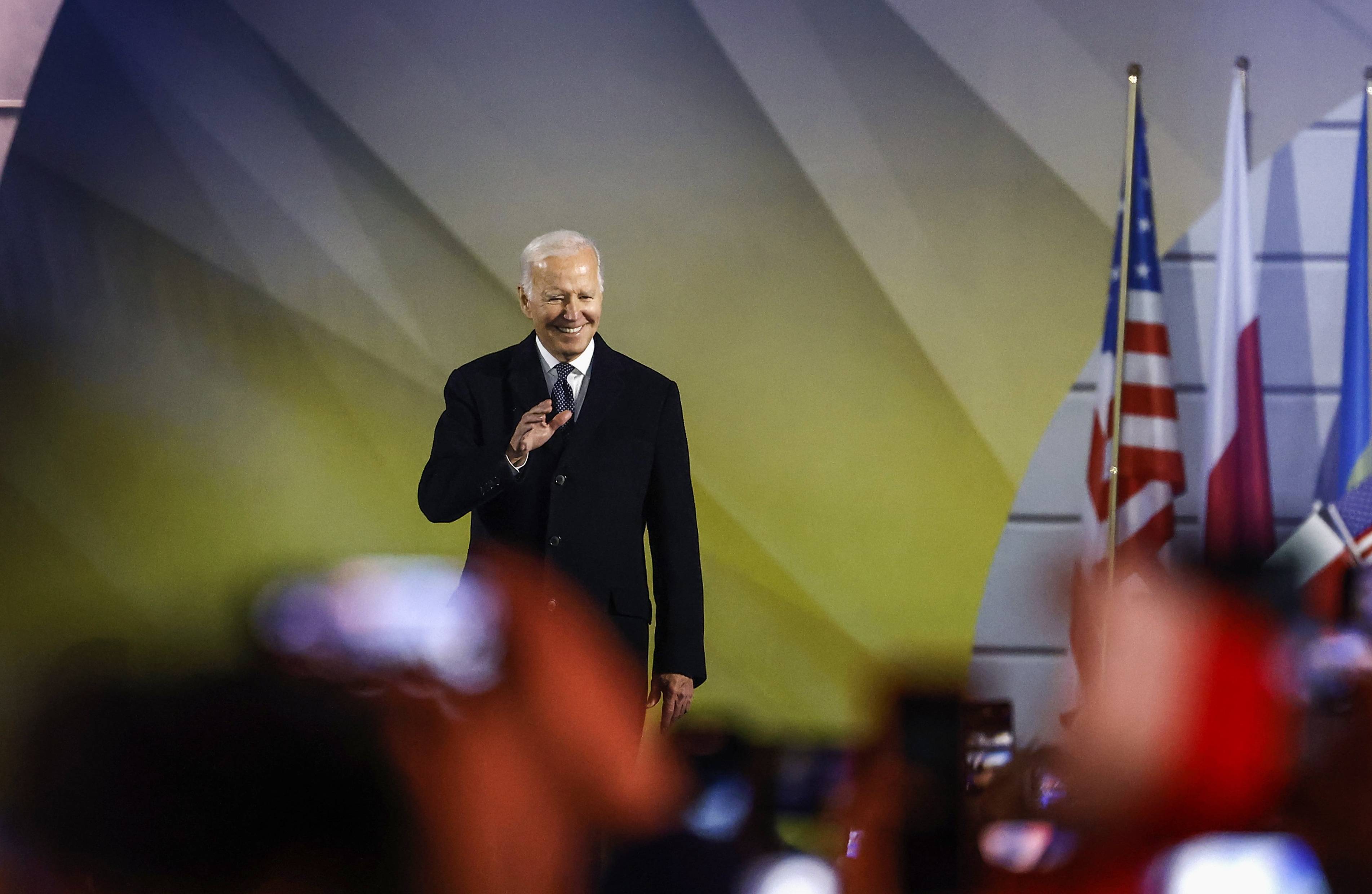 Joe Biden na tle flag Polski, Ukrainy i USA. Jest uśmiechnięty, macha do tłumu w Warszawie