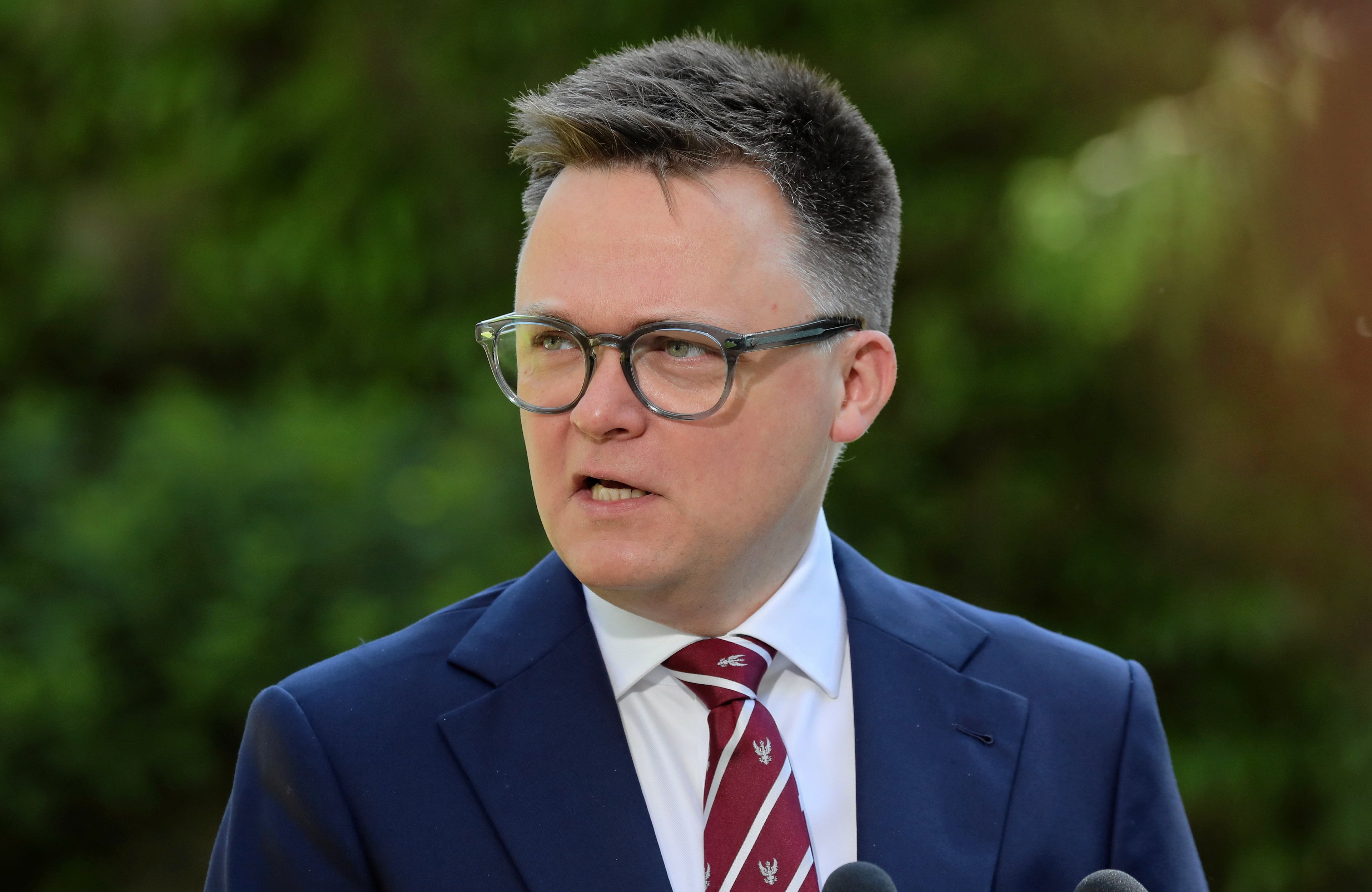 Szymon Hołownia w garniturze na tle zieleni. Patrzy w prawo i przemawia do mikrofonu.