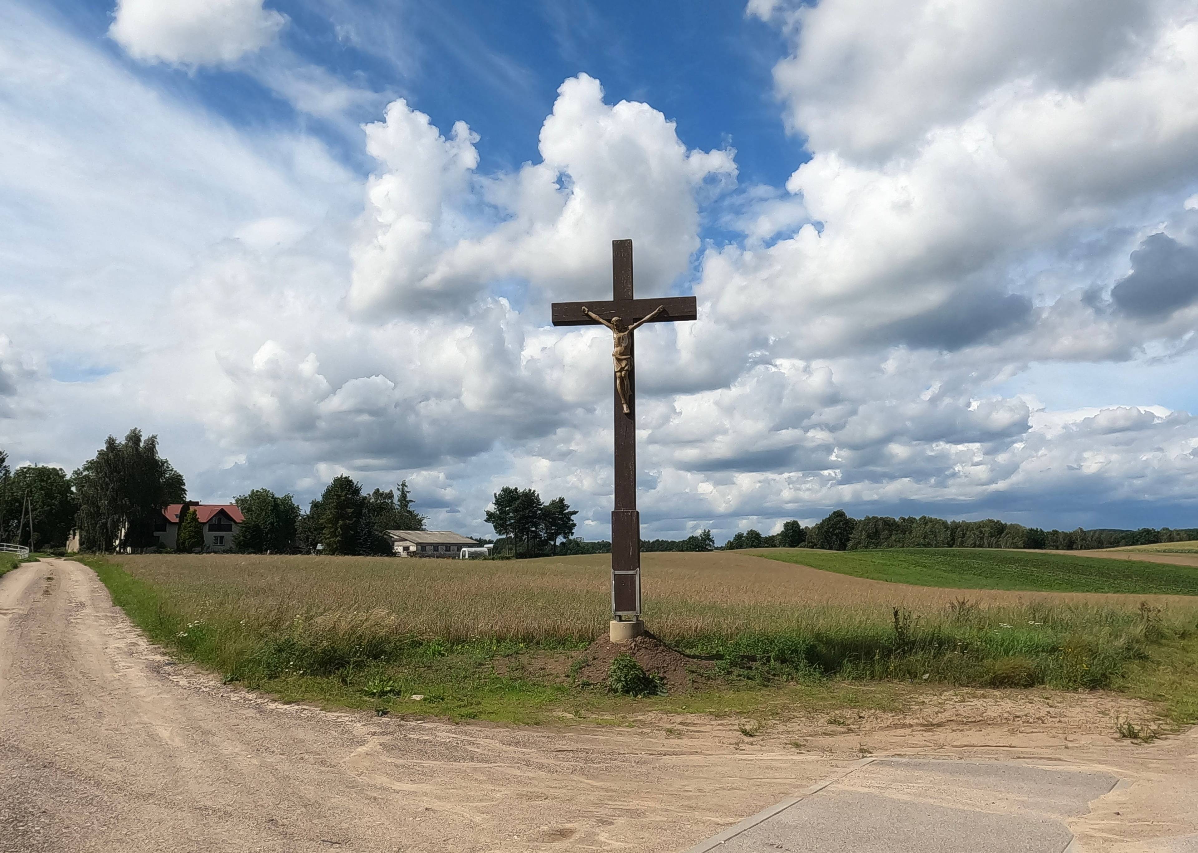 drewniany krzyż na rozstaju dróg, w oddali zabudowania