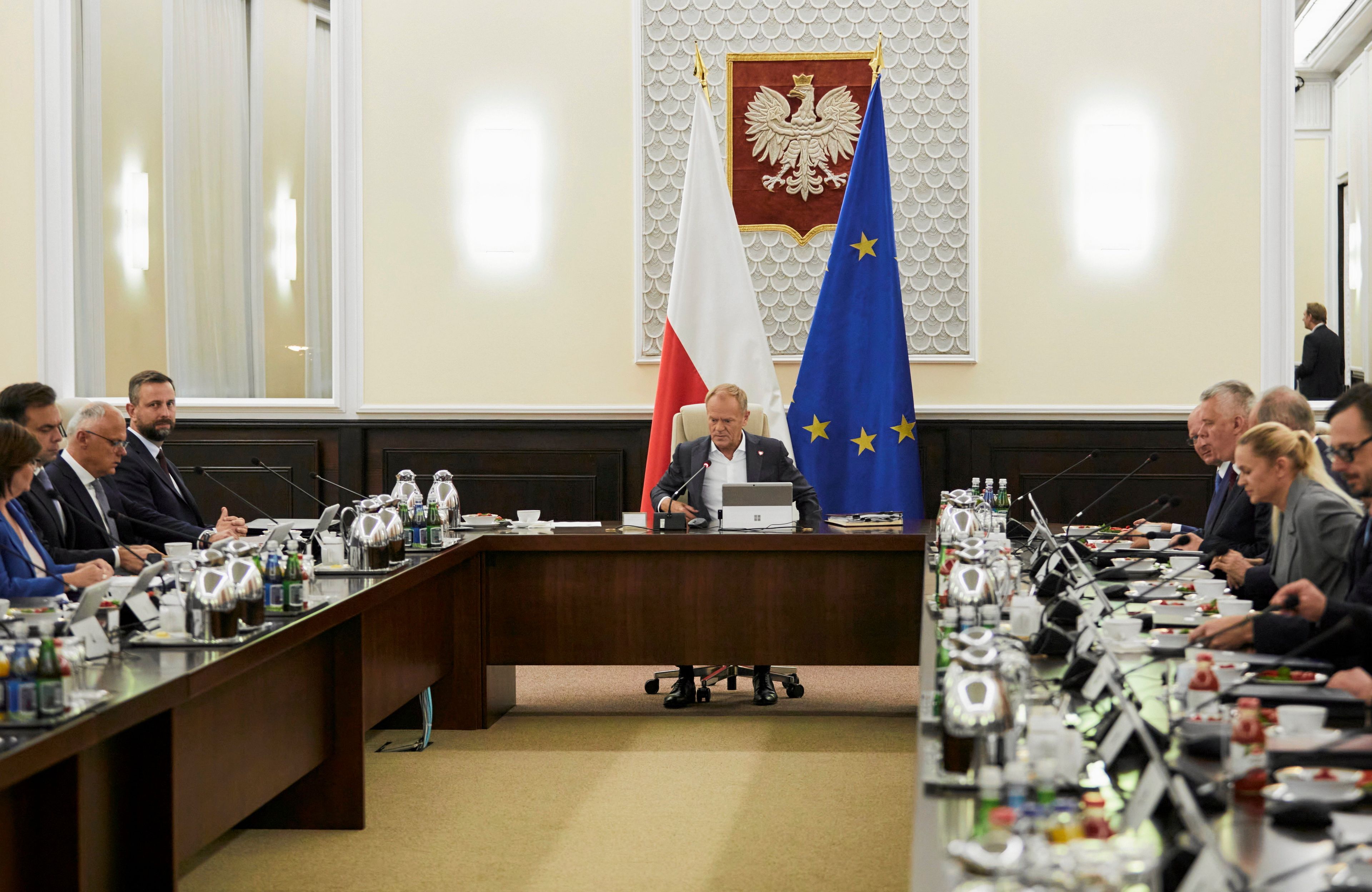 Za kwadratowym stołem siedzą ministrowie, w szczycie premier, za nim flagi UE i Polski