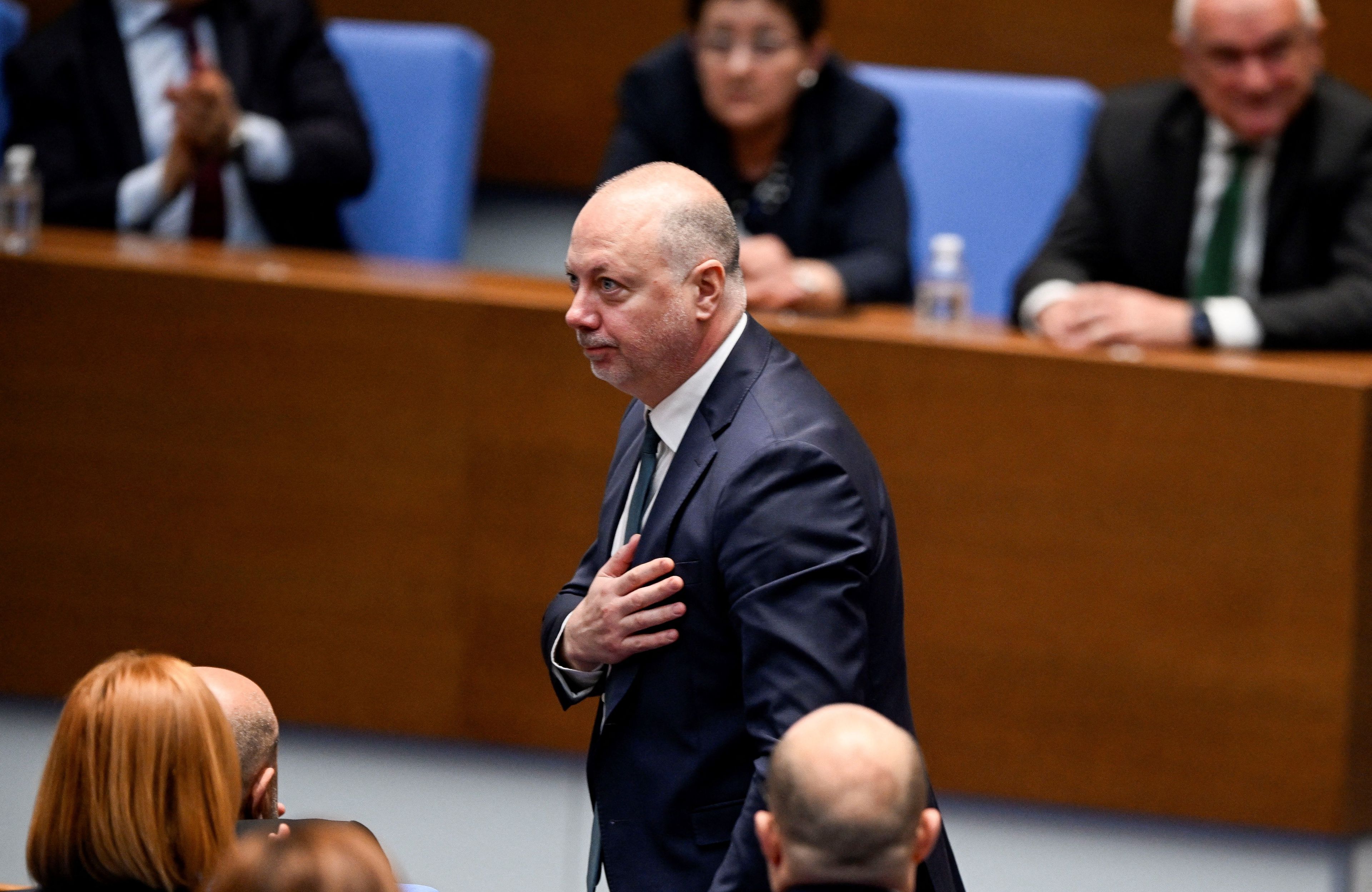 Mężczyzna w ciemnym garniturze stoi w parlamentarnych ławach, trzymając prawą rękę na sercu