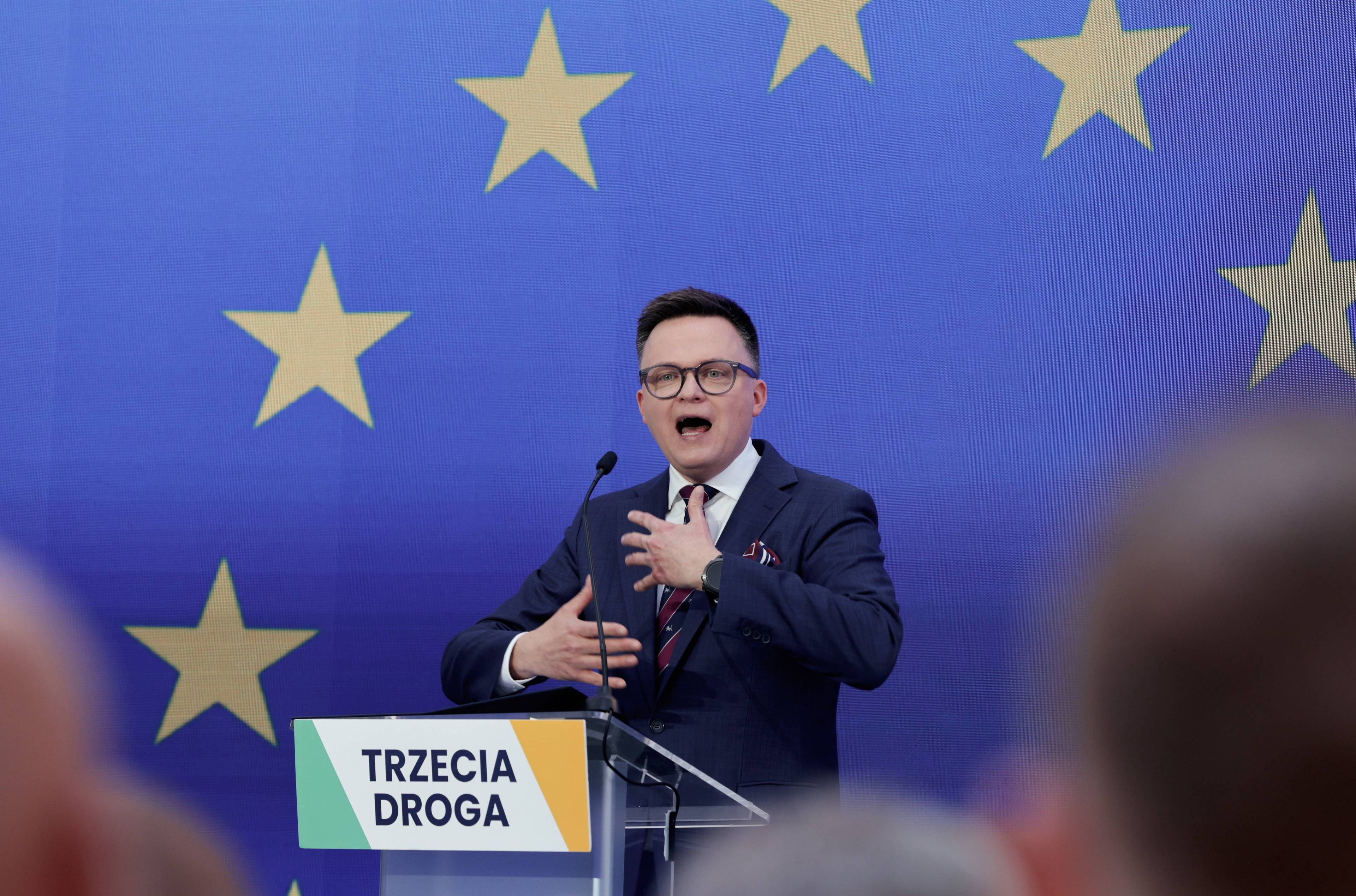 Szymon Hołownia przemawia na konwencji przed eurowyborami. Stoi za mównicą z napisem "Trzecia Droga", za nim niebieskie tło i gwiazdki z flagi Unii Europejskiej. Wybory europejskie