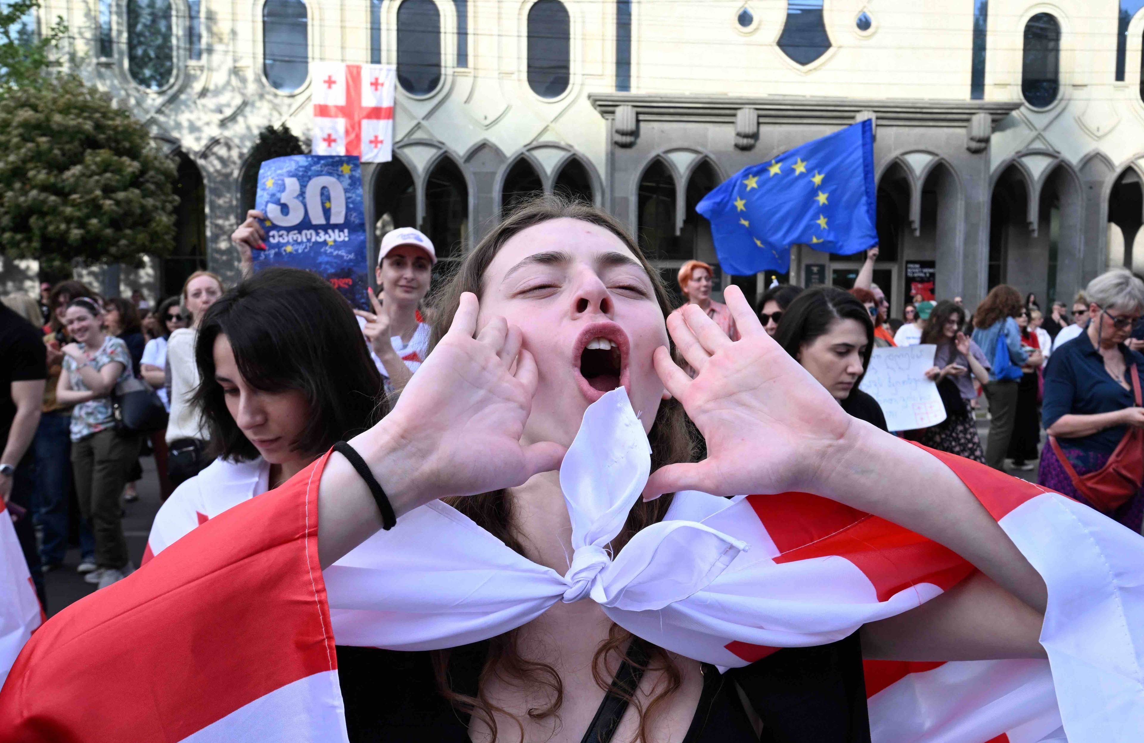 Uczestniczka demonstracji krzyczy, przykładając dłonie do ust, na ramionach ma gruzińską flagę narodową, w tle inni demonstranci z flagami gruzińskimi i UE. Gruzja