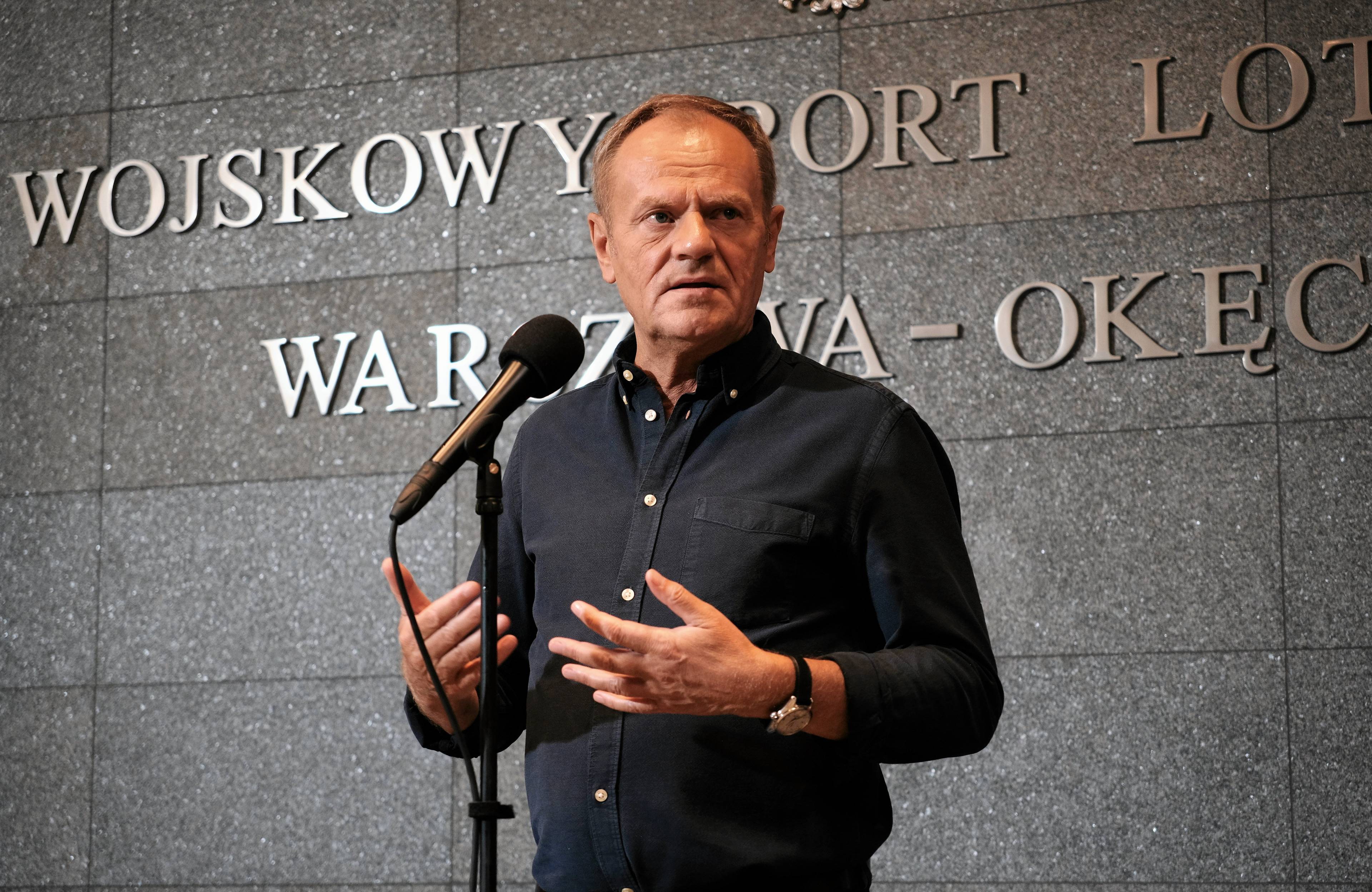 Premier Donald Tusk przed mikforonem w ciemnej koszuli na tle szarej ściany
