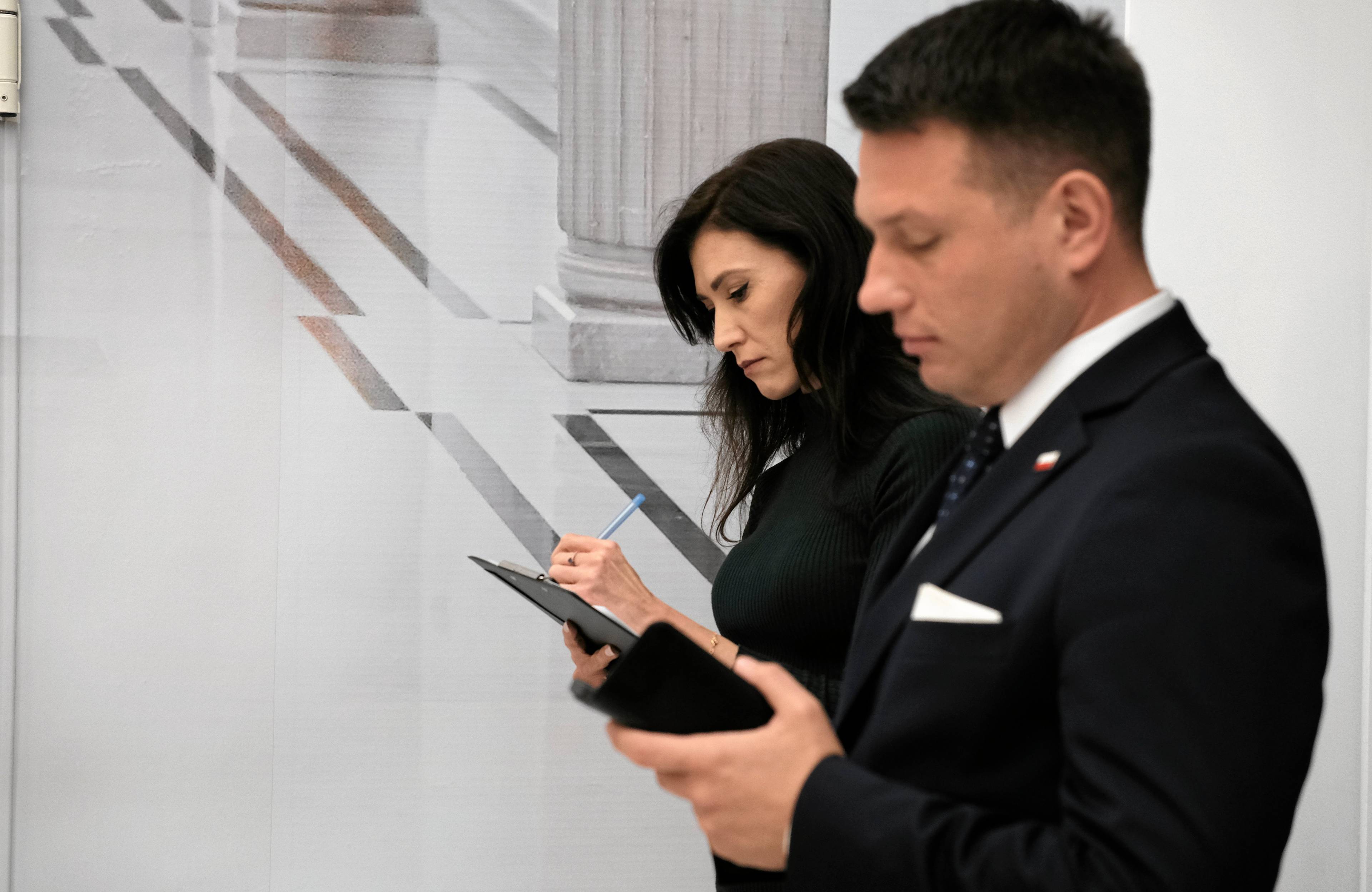 Mężczyzna i kobieta w holu Sejmu. Stoją, czytając coś w telefonach komórkowych. Oboje w strojach oficjalnych