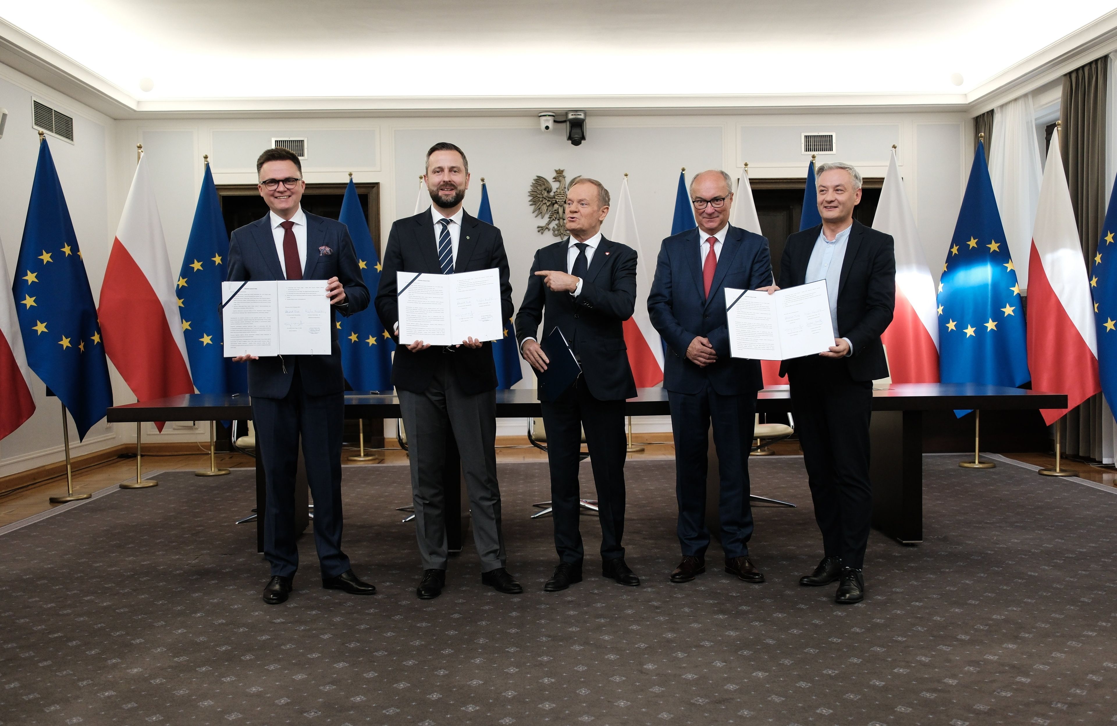 Liderzy Trzeciej Drogi, KO i Nowej Lewicy stoją obok siebie, trzymając podpisaną umowę koalicyjną, w tle flagi Polski i Unii Europejskiej
