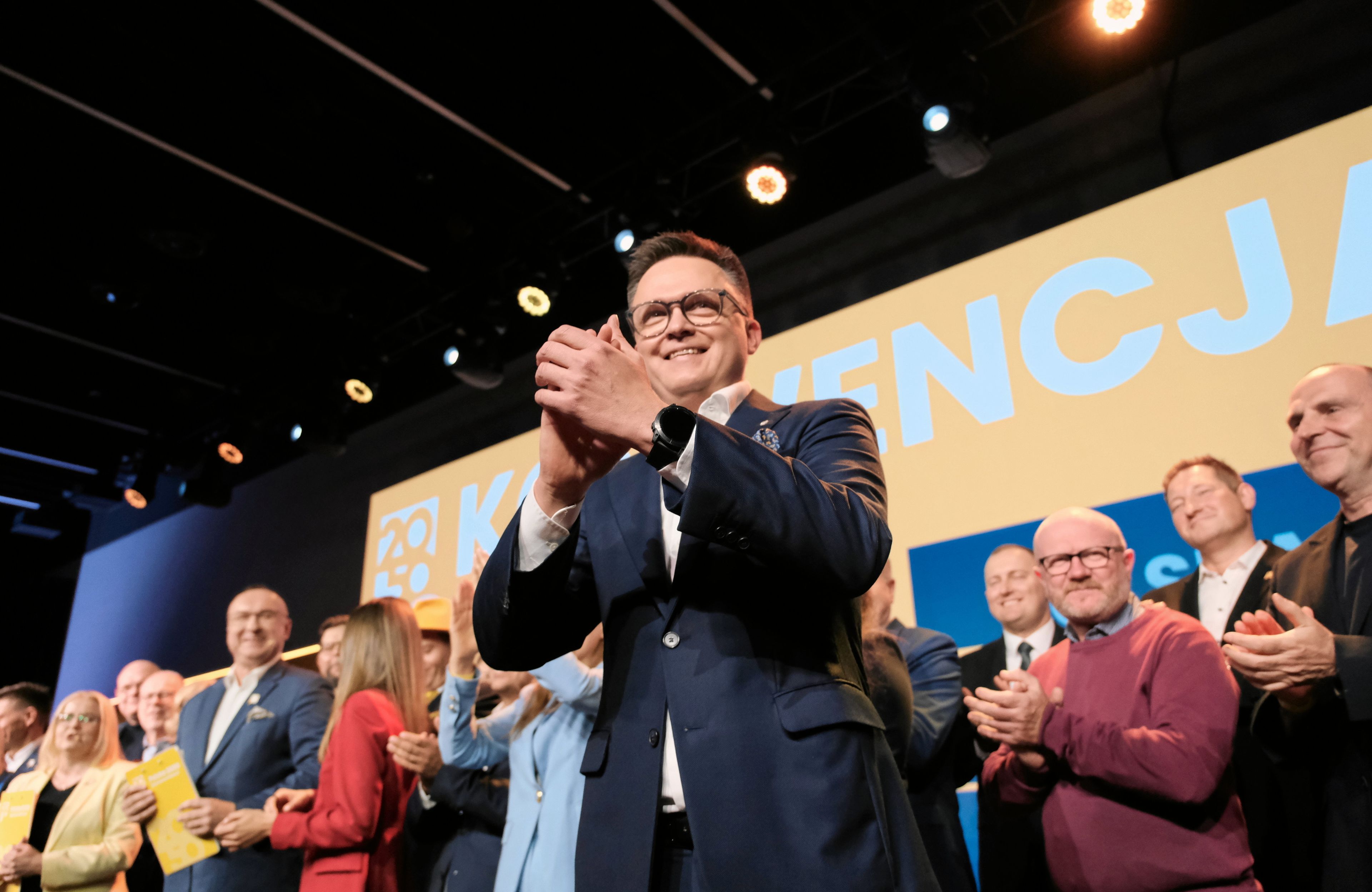 Szymon Hołownia klaszcze w dłonie na tle białego napisu "Konwencja" na żółtym tle