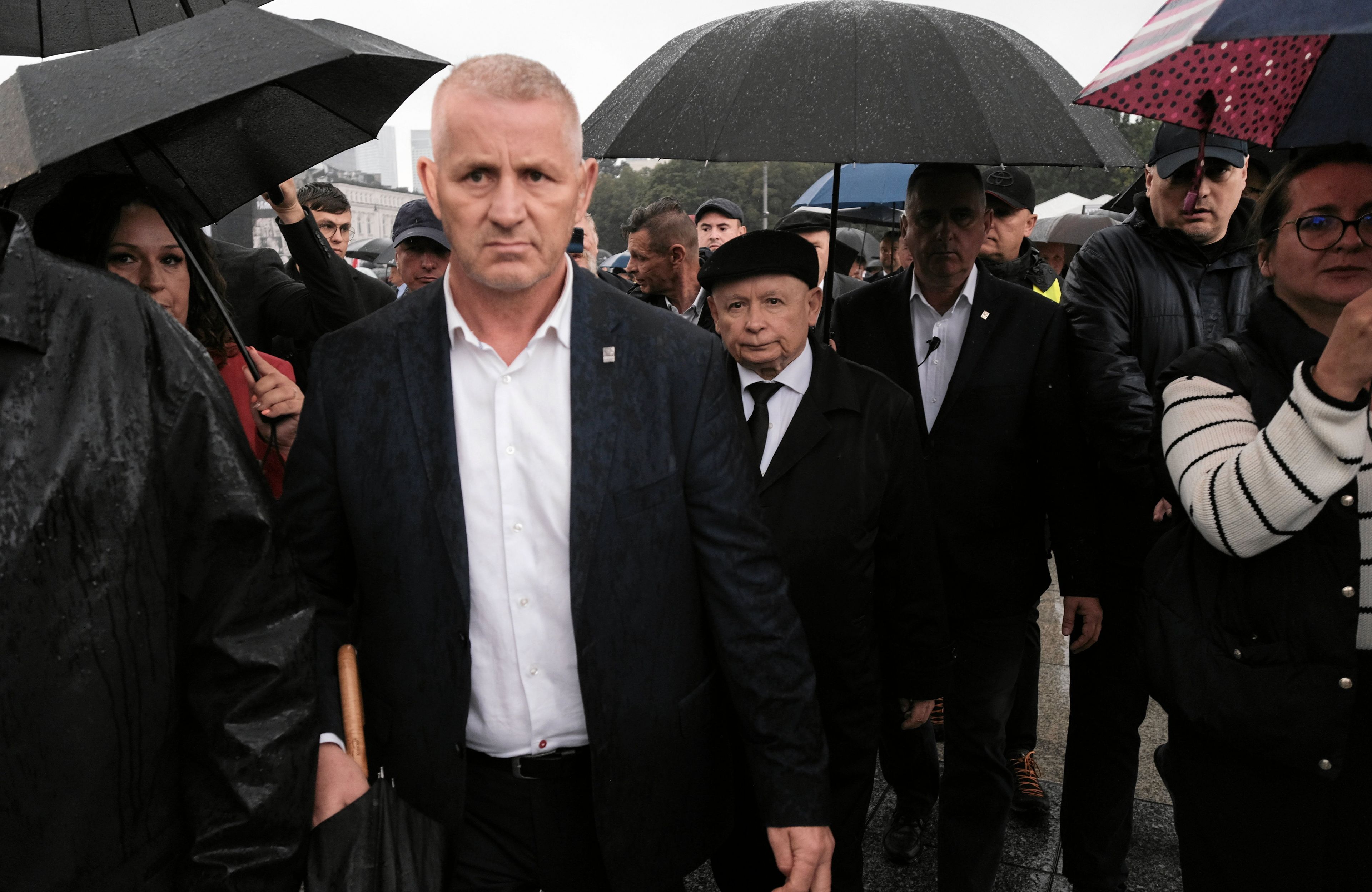 Jarosław Kaczyński w otoczeniu ochroniarzy na placu Piłsudskiego podczas miesięcznicy smoleńskiej. Pada deszcz, ochroniarze niosą nad Kaczyńskim otwarte parasole