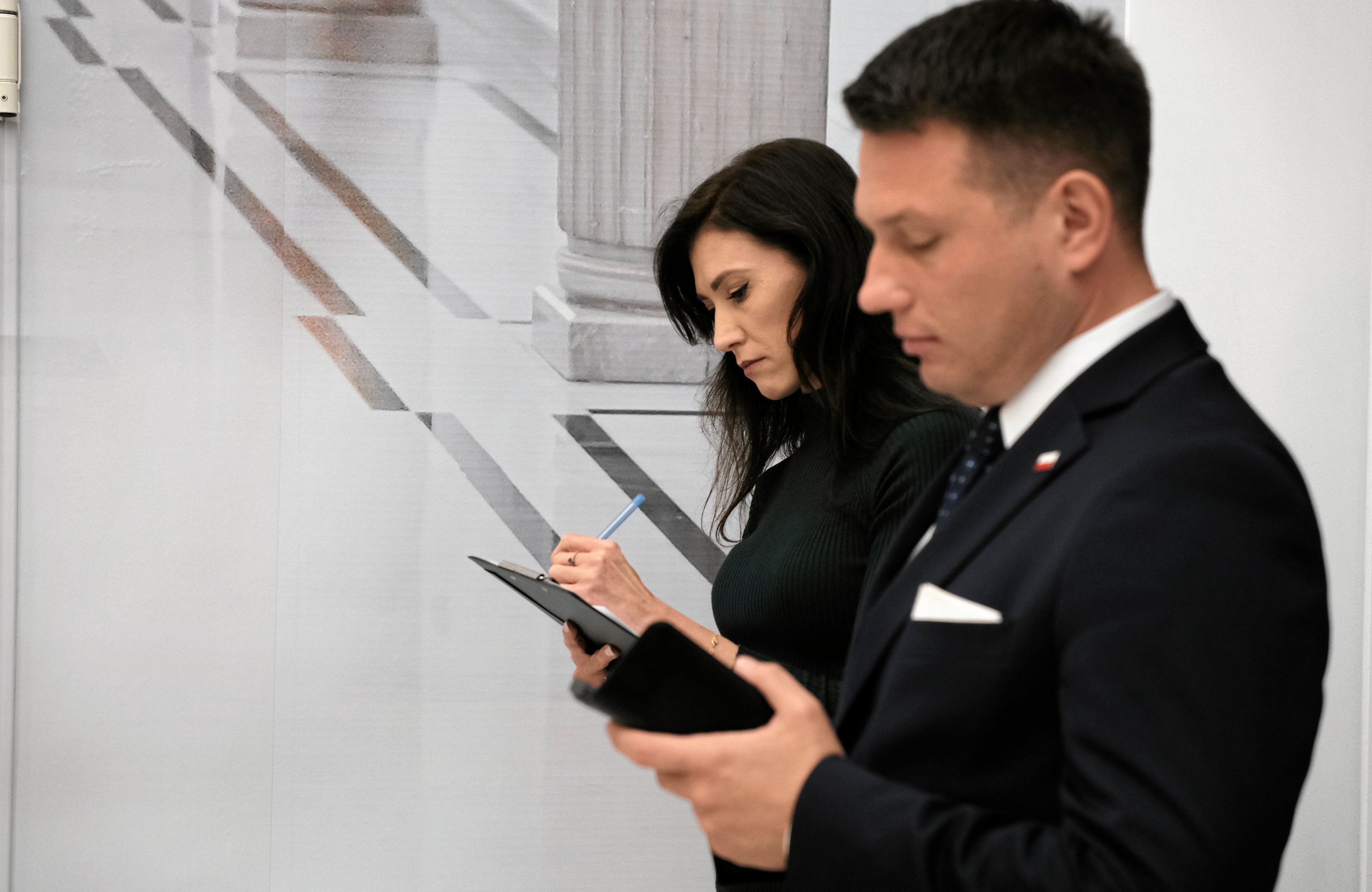 Mężczyzna i kobieta w holu Sejmu. Stoją, czytając coś w telefonach komórkowych. Oboje w strojach oficjalnych