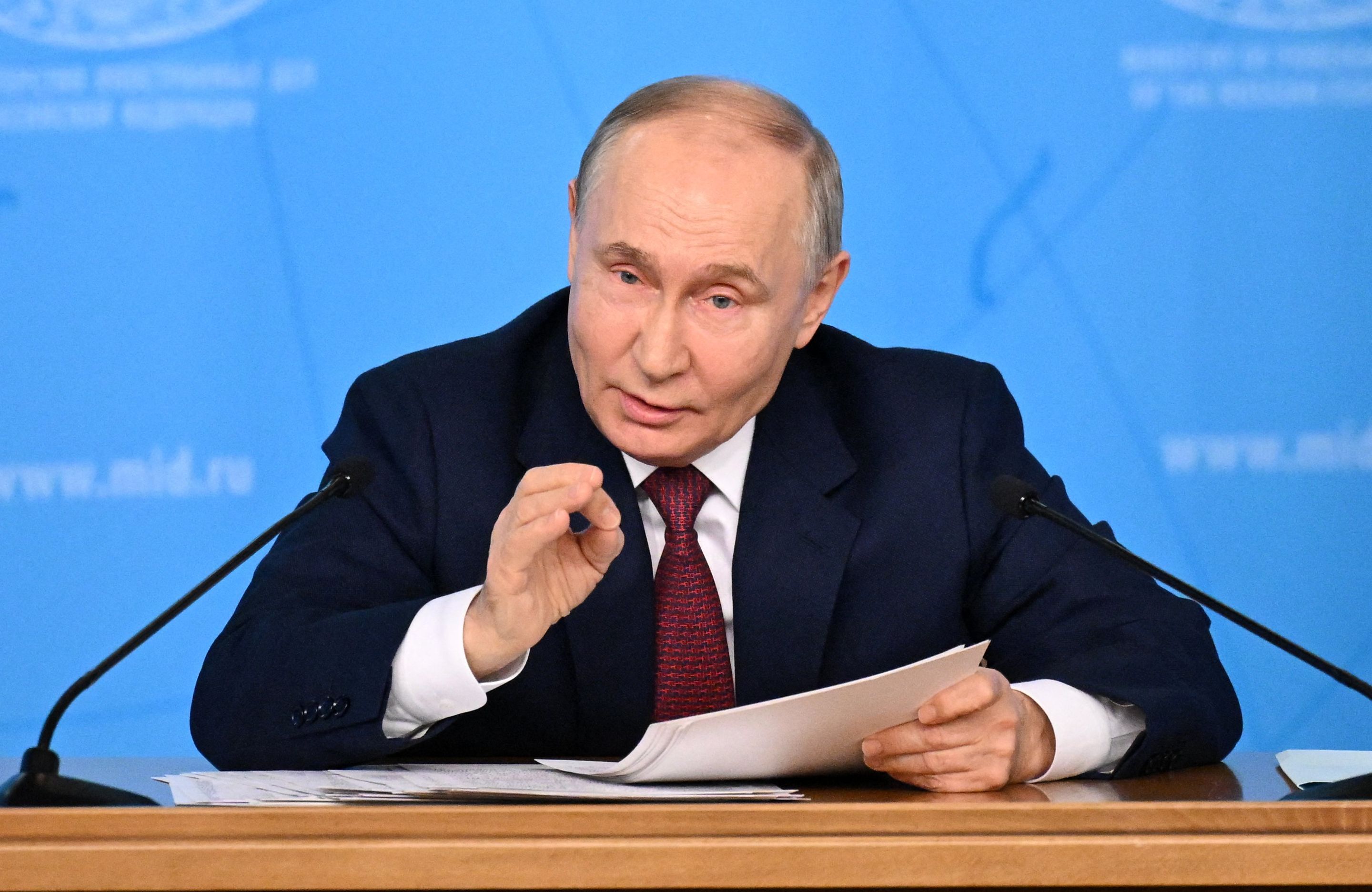 Władimir Putin. Łysiejący, krępy mężczyzna w garniturze i białej koszuli, siedzi przy stole, gestykuluje jedną dłonią.